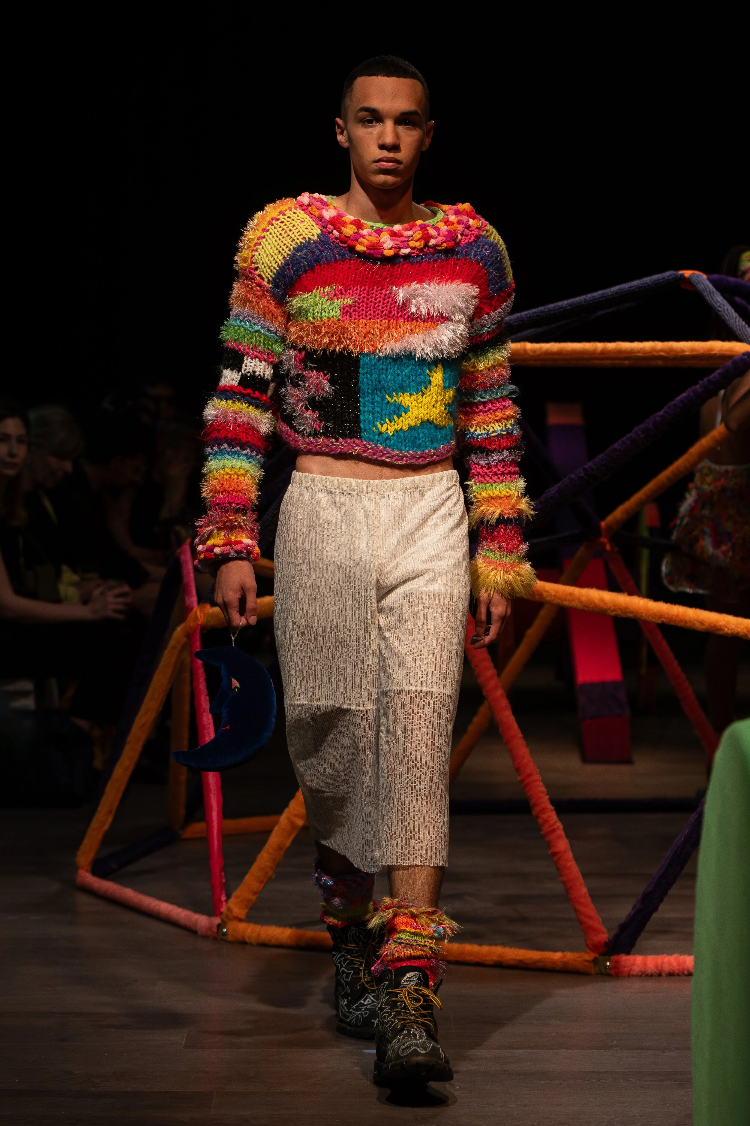 Model walking on runway in colorful, eclectic sweater with various textures, patterns, and a yellow star on the front, paired with beige pants, colorful socks, and combat boots, during fashion show.