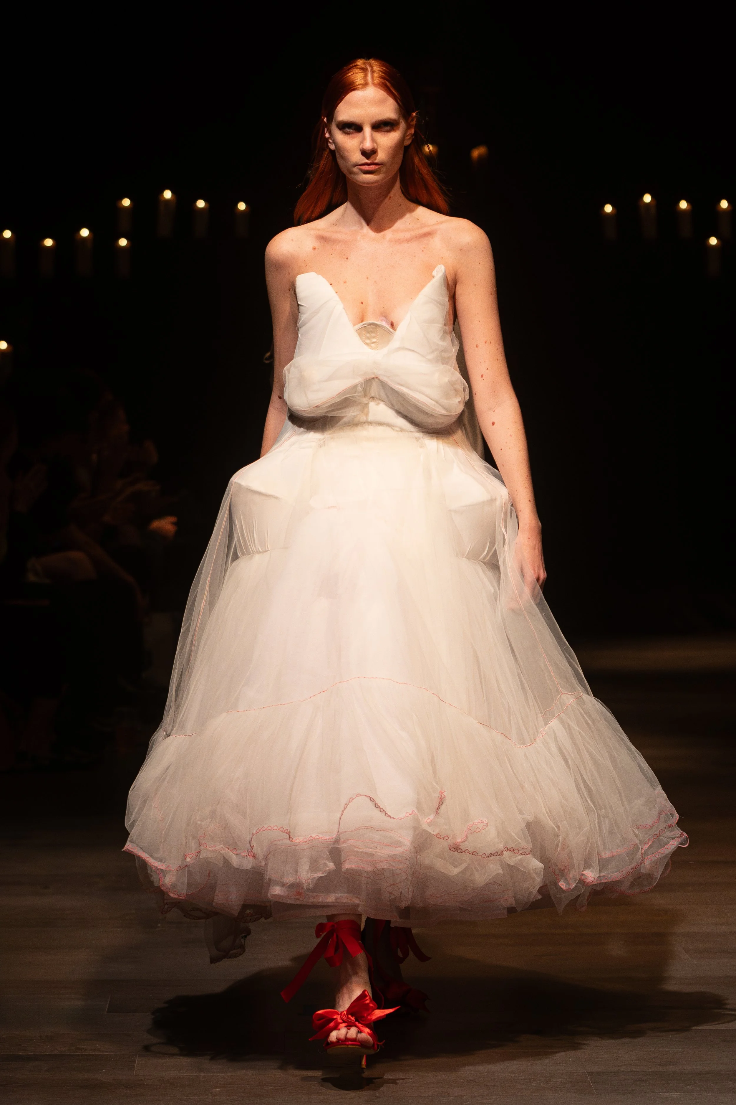 A woman with red hair walking down a runway in a strapless, puffy white dress with red shoes and ribbons.
