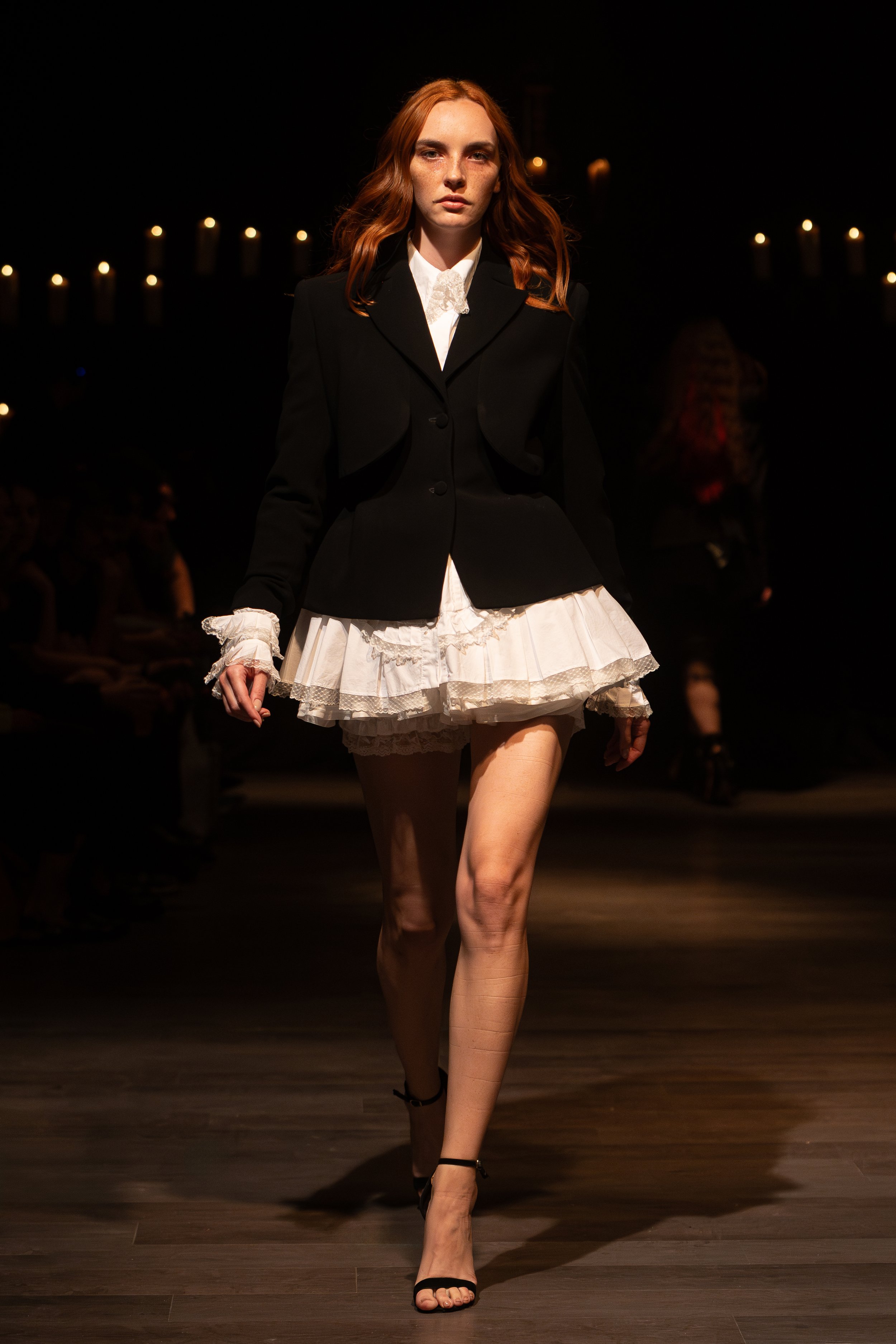 A slender woman with long legs, wearing a black blazer with a white ruffled blouse underneath, walking on a runway with a dark background and candles. She has wavy red hair and a confident expression.