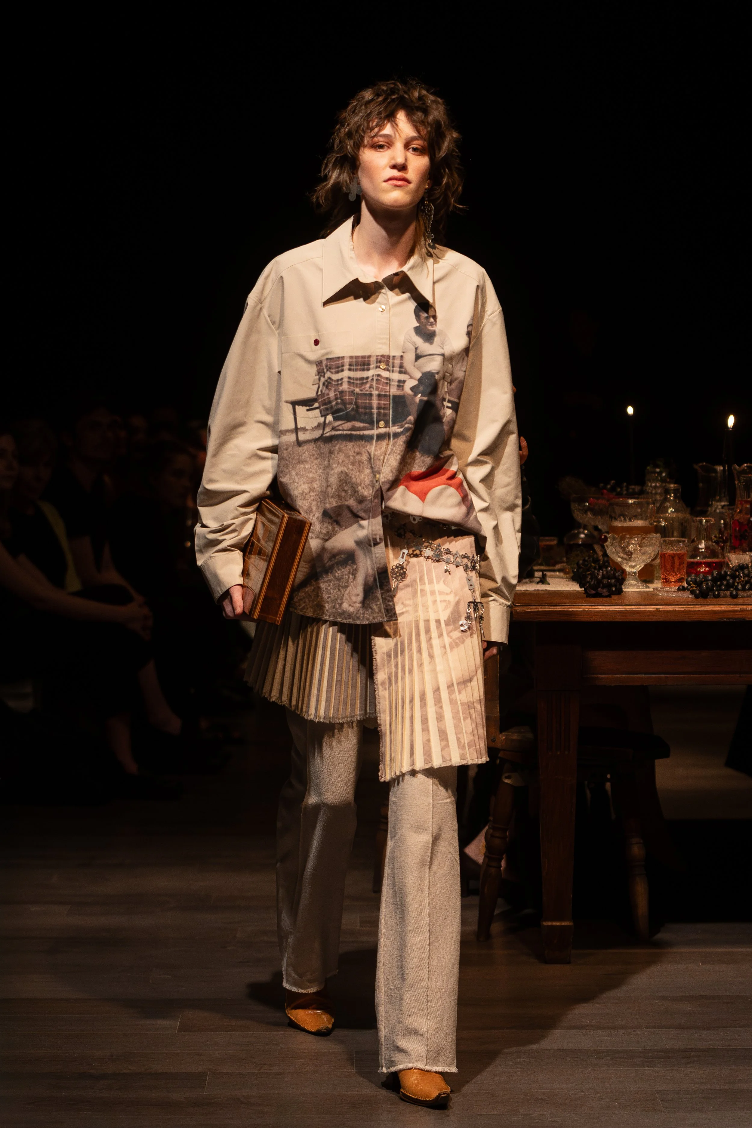 A woman walking on a runway during a fashion show, wearing a beige and patterned coat over a pleated skirt, with dark styled hair and earrings, set against a dark background with an audience and a table with glassware and fruits.