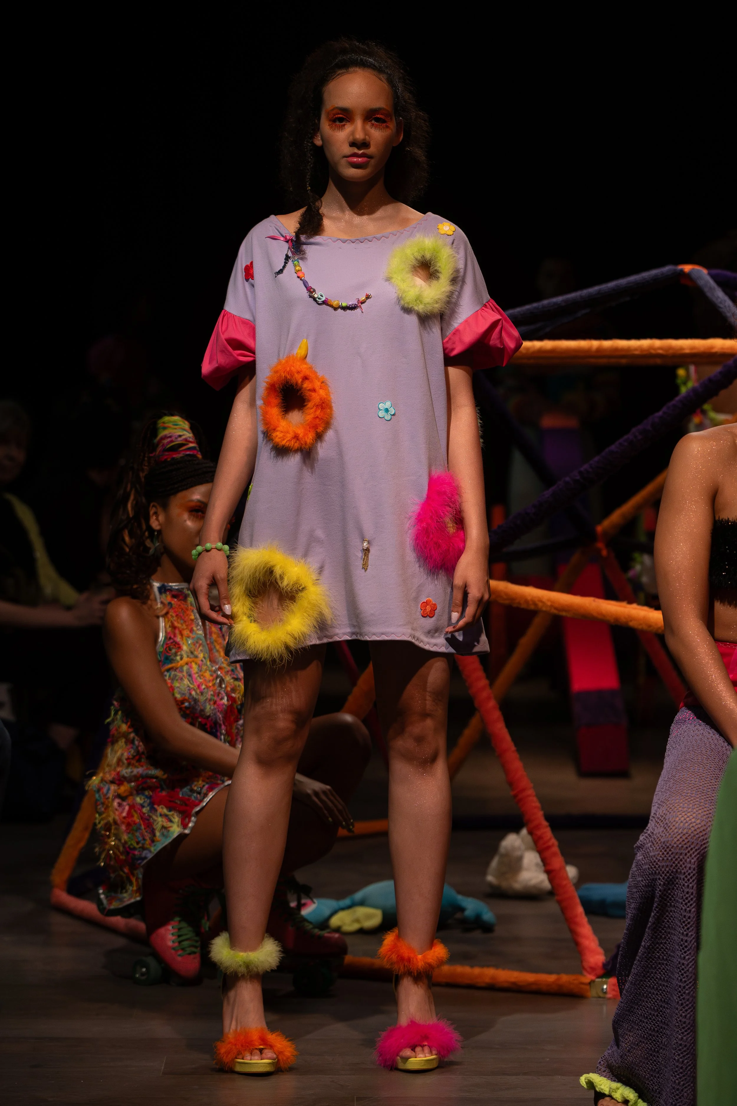 Fashion model standing on runway wearing colorful, fuzzy accessories and a loose, pastel-colored dress during a fashion show.