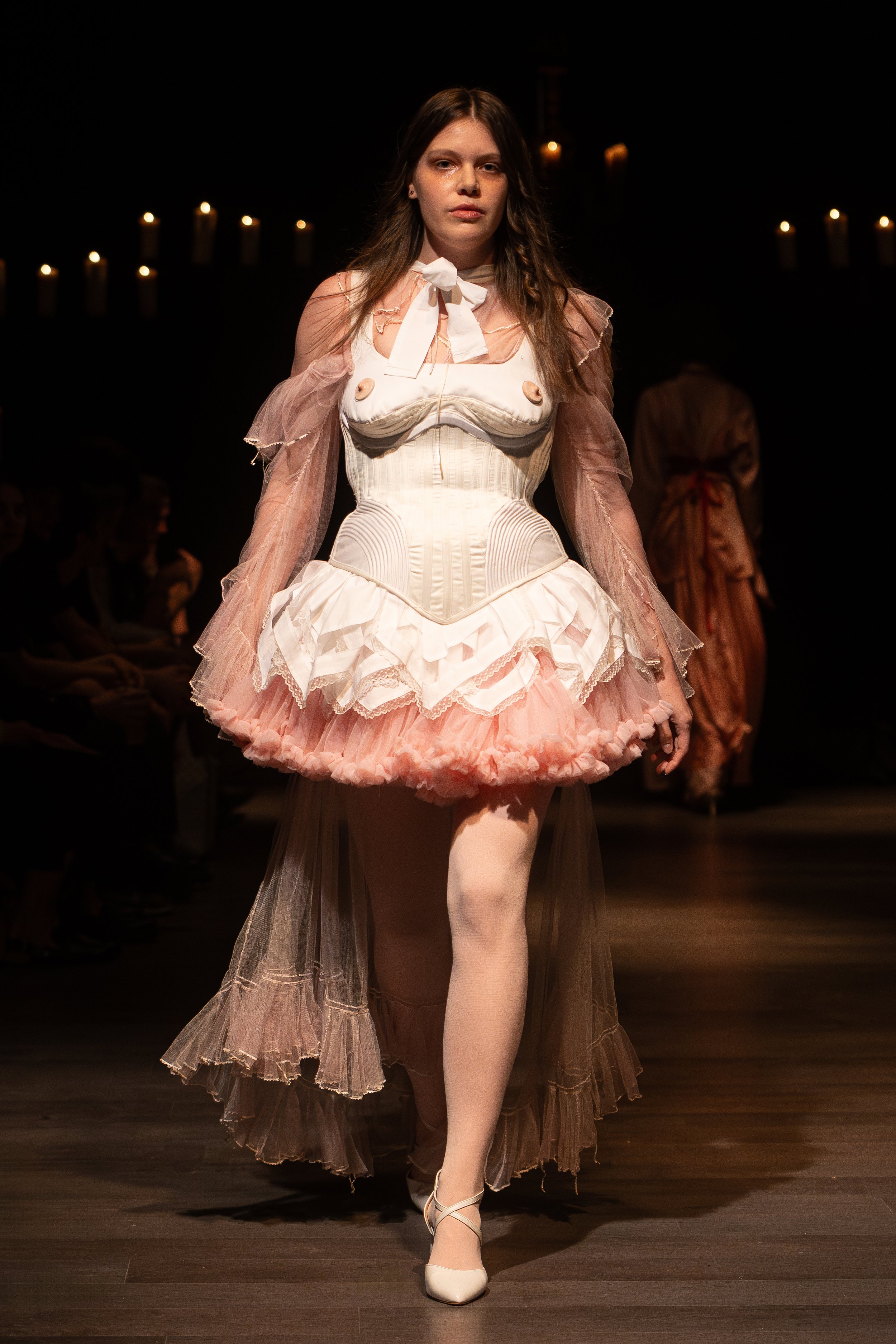 A female model walking on a runway wearing a Victorian-style dress with a corset and ruffled skirt, in front of a dark background with candles.