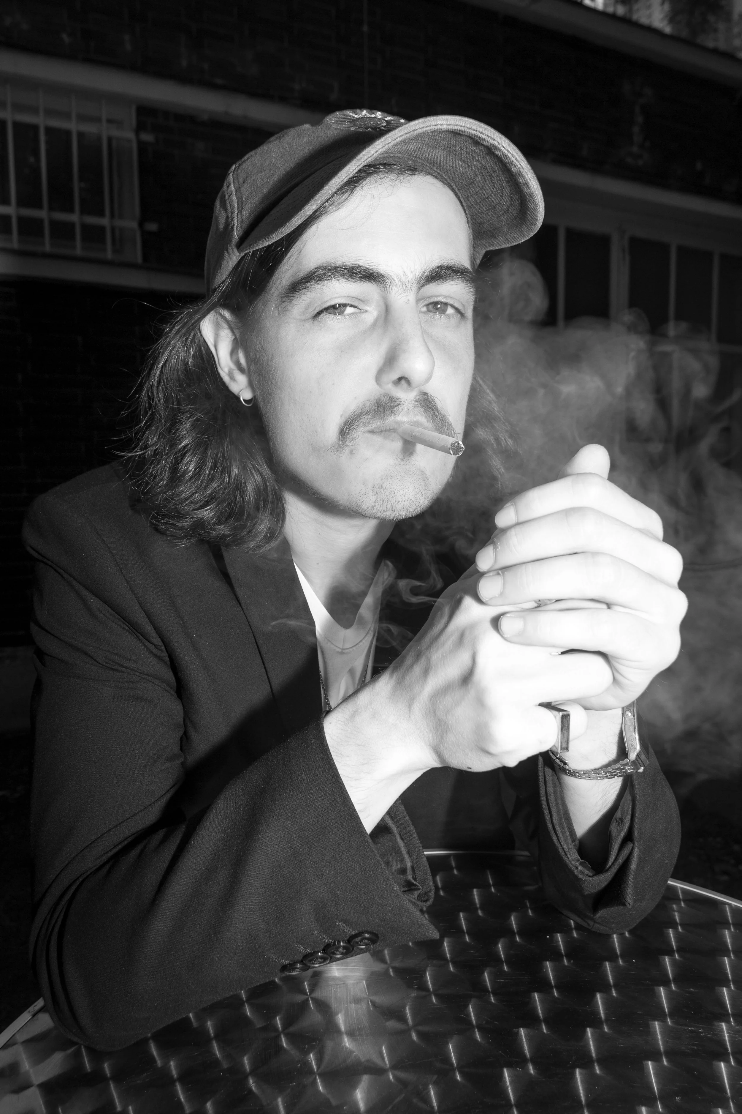 Black and white photo of a Olasz Renáta  with long hair, a mustache, and beard, wearing a cap, smoking a cigarette, and resting his hands together on a metallic table with a textured pattern photographed by Bede Kincső Hilda at @notallowedshows