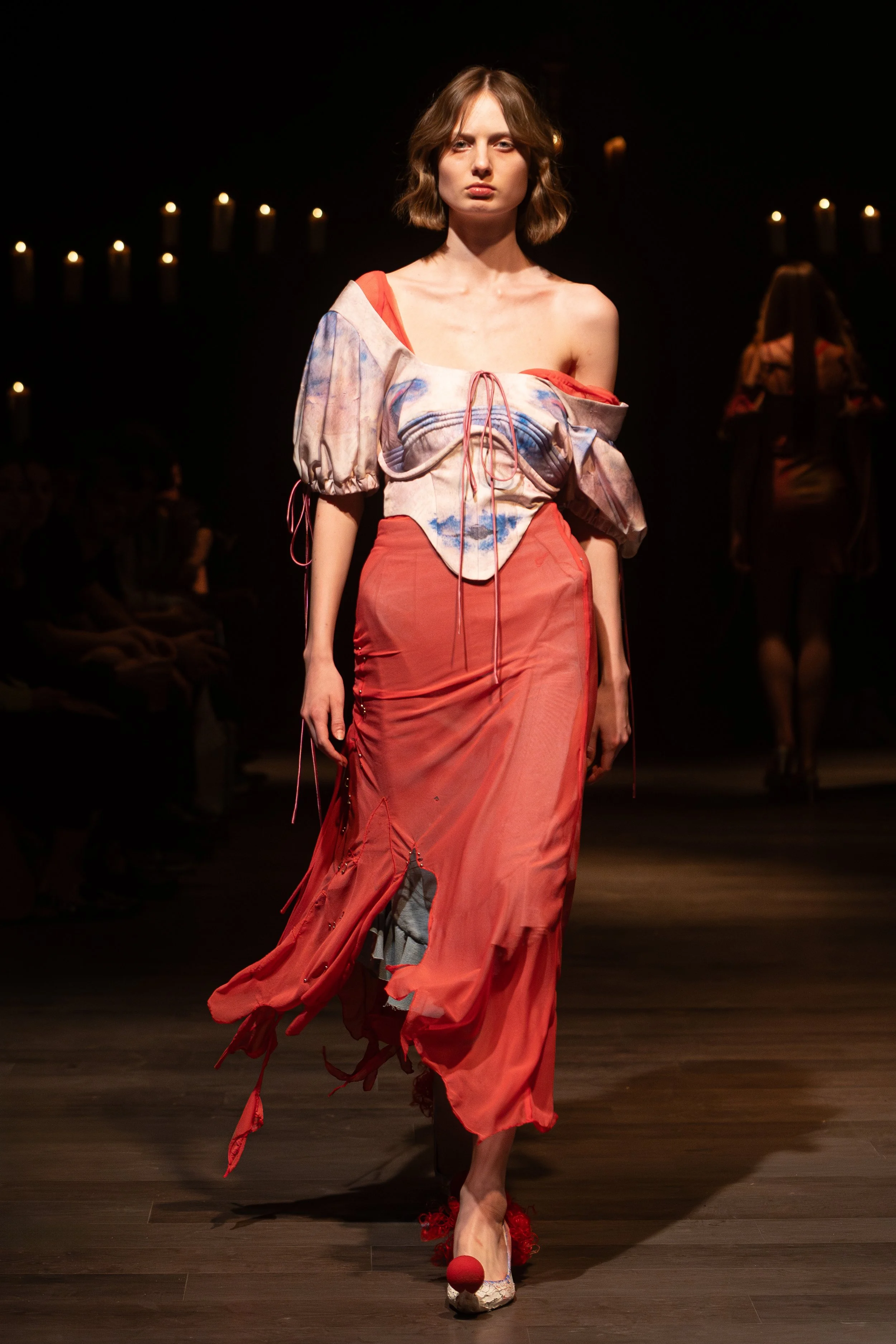 A female model walks down a runway wearing a red dress with layered, ruffled details, and unique, artistic footwear with large bow accents. The background is dark with soft lighting and candles, focusing attention on the model and her outfit.
