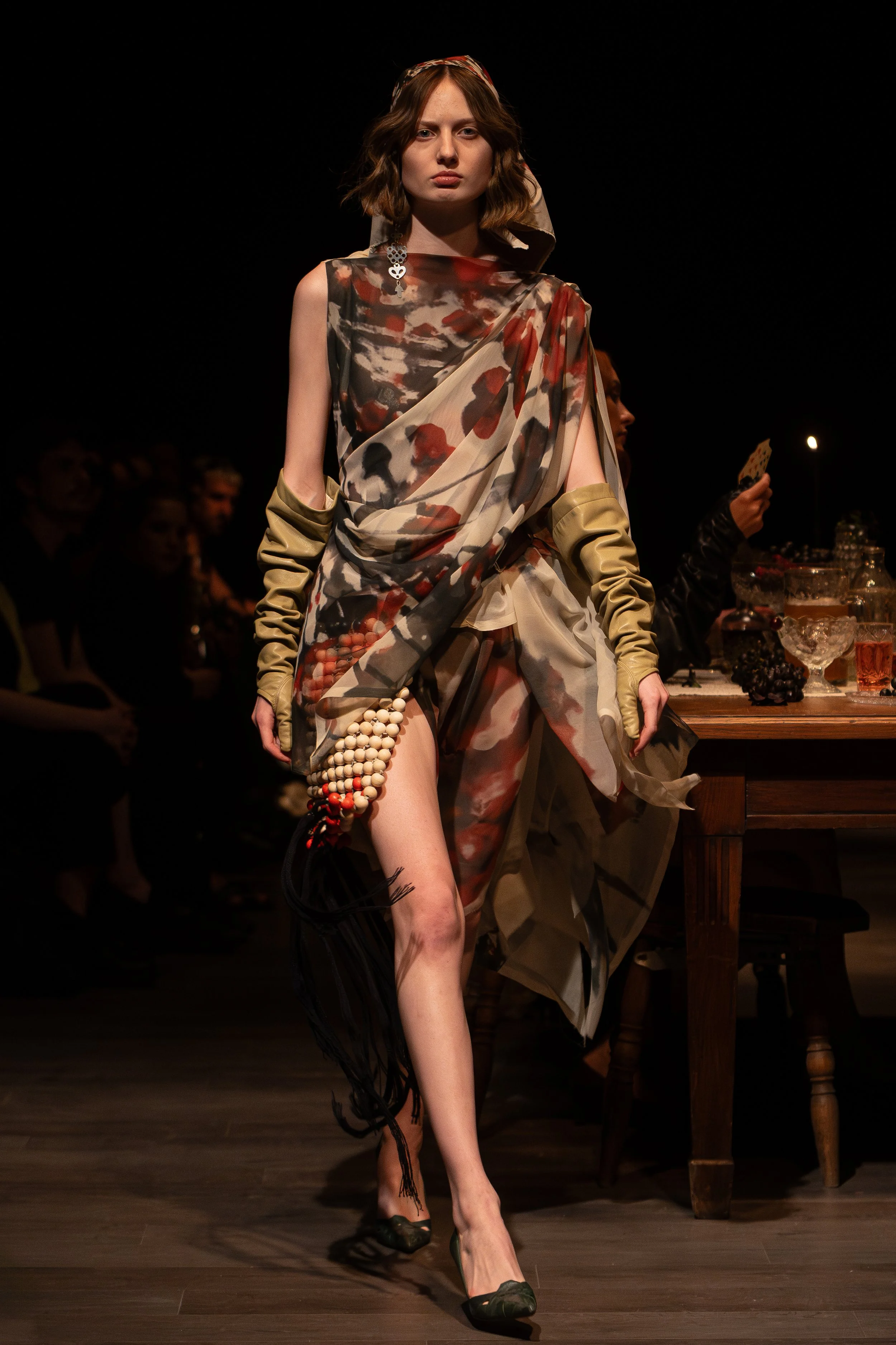 Model walking on runway wearing patterned dress with layered fabric, beige sleeves, and black shoes at fashion show.
