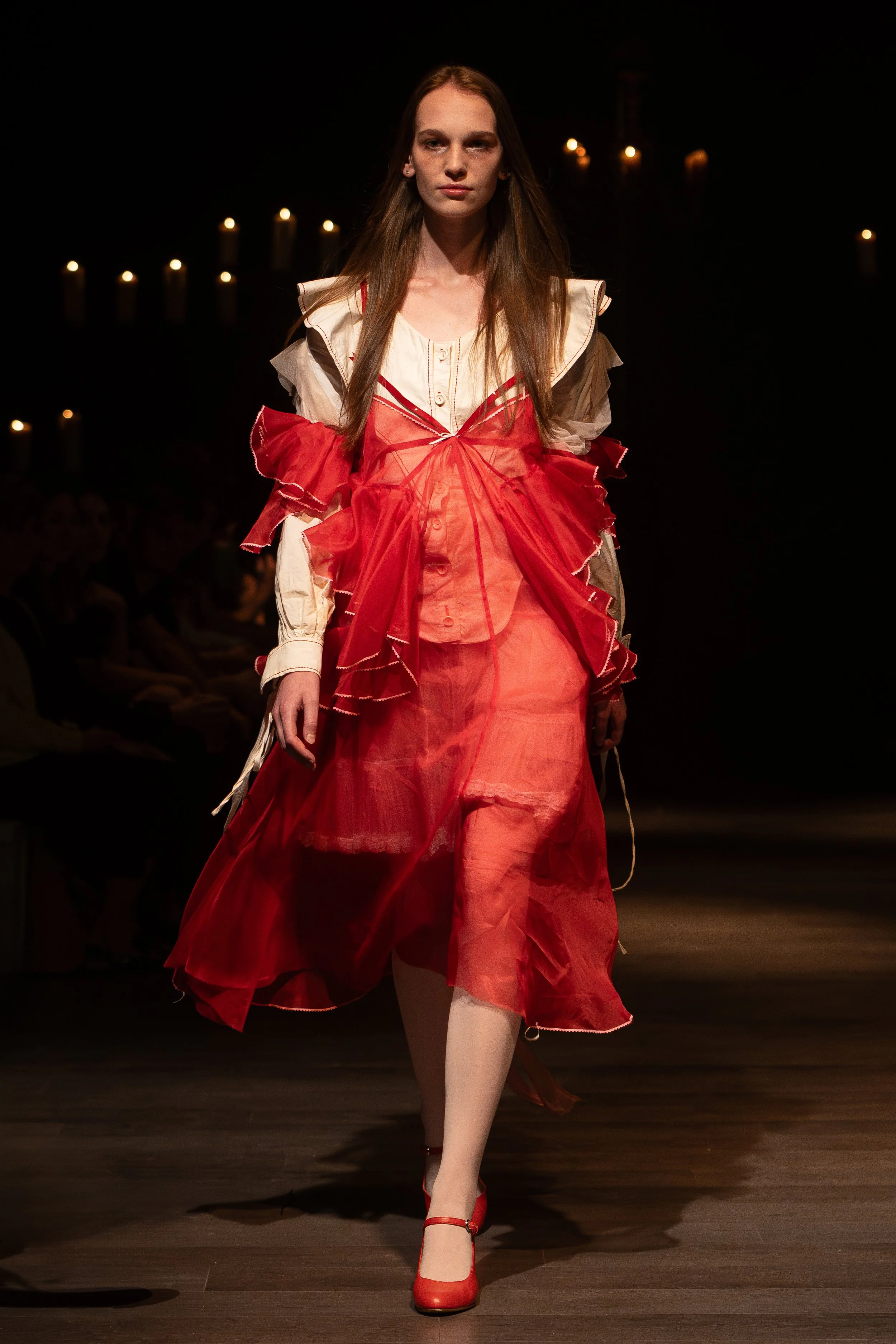 A female model walking down a runway in a fashion show, wearing a layered, flowing dress with red, beige, and brown colors, and red high-heeled shoes, with candles in the background.