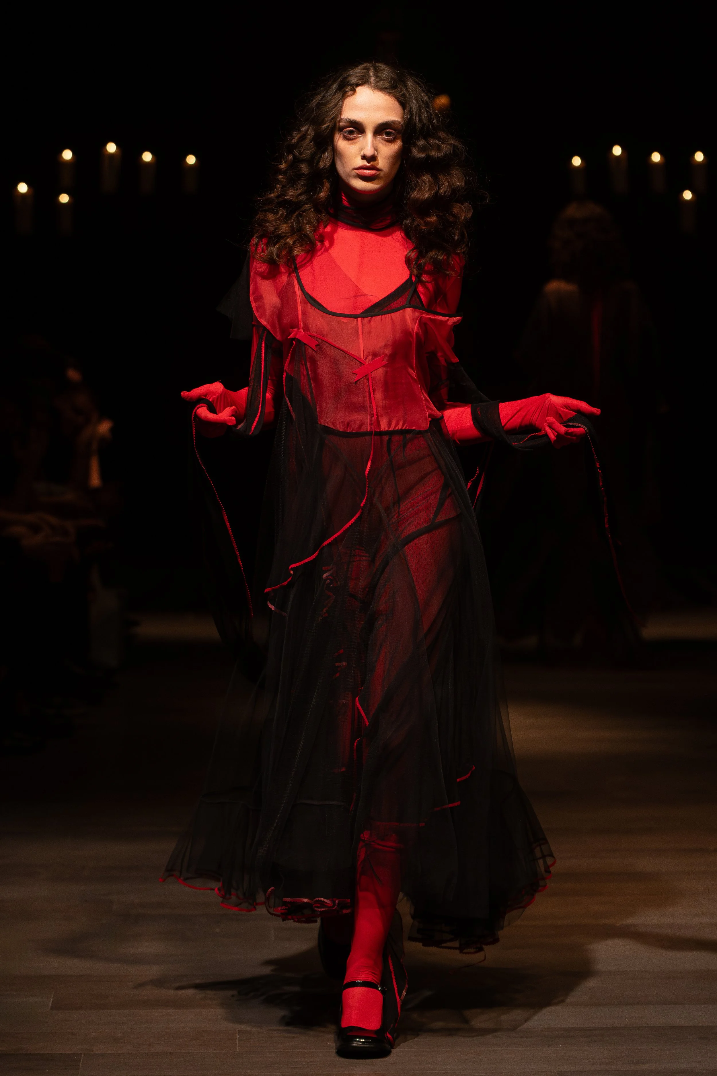 Model walking on runway wearing sheer red and black layered dress with red gloves and red shoes, dark background with candles.