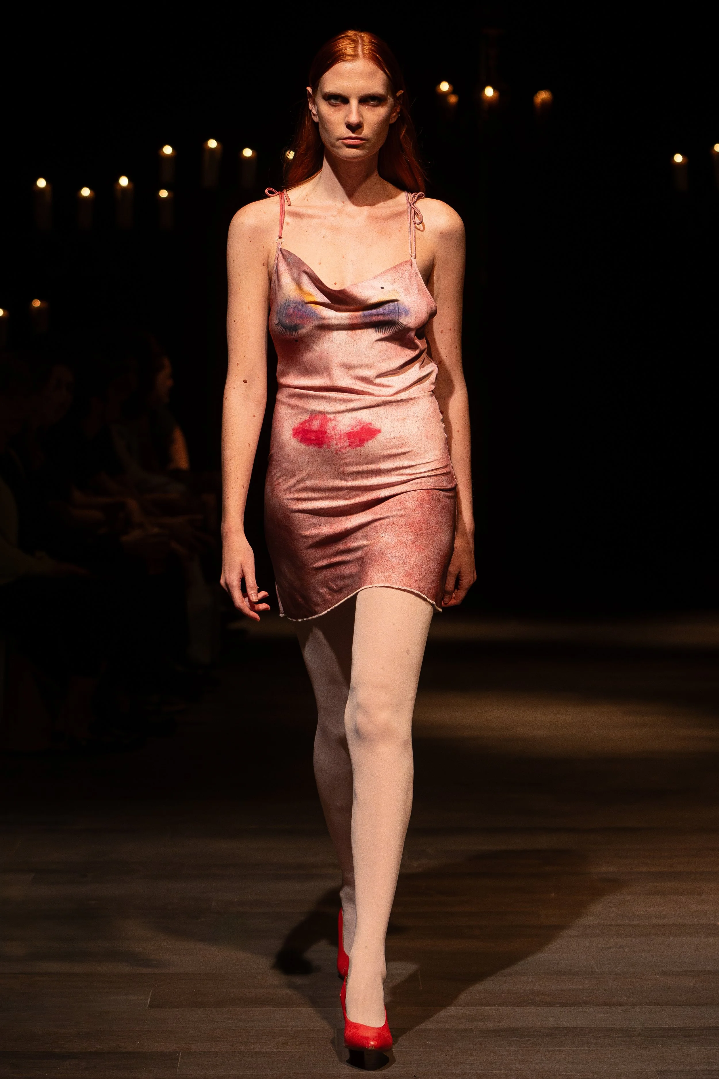 A female model walking down a runway wearing a pink satin dress with lipstick print and red high heels, with candles in the background.