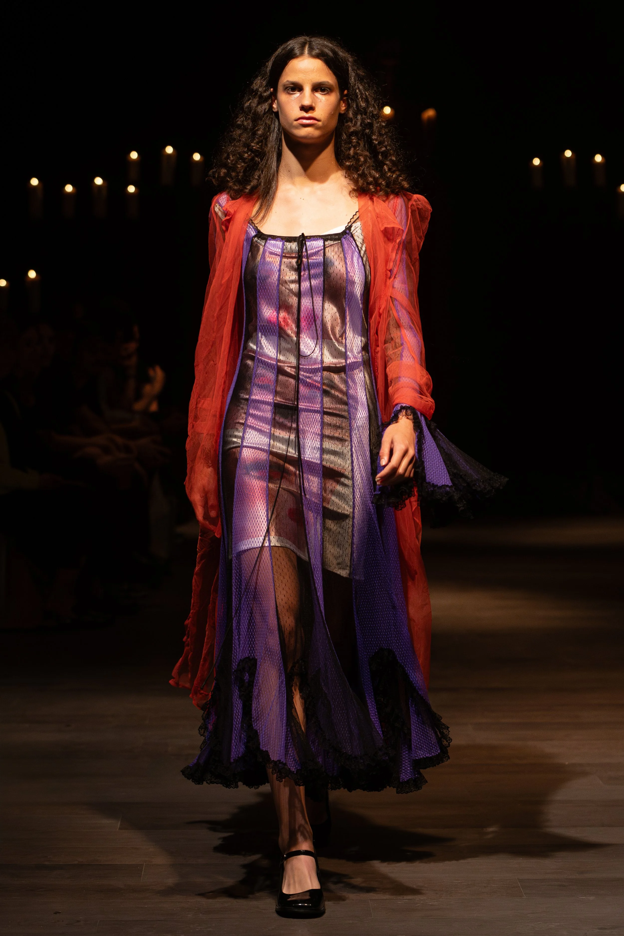 A woman walking down a runway at a fashion show, wearing a colorful layered dress with lace and mesh details and a sheer red overlay. She has dark curly hair and stiletto heels, with audience members visible in the background.