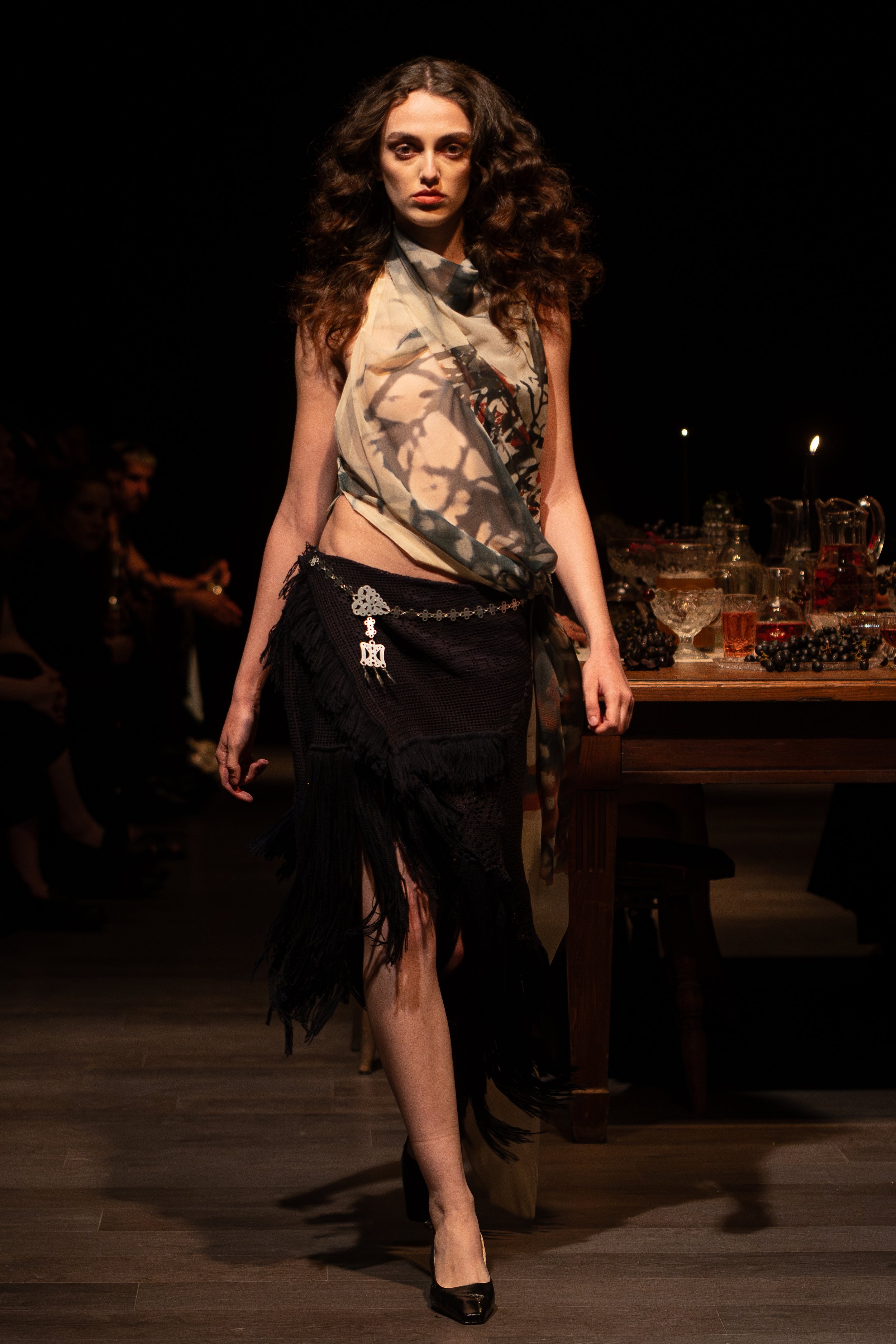 A woman with curly hair wearing a patterned scarf, a black fringed skirt, and black high heels, walking confidently on a runway with a dark background and tables with drinks and people in the background.