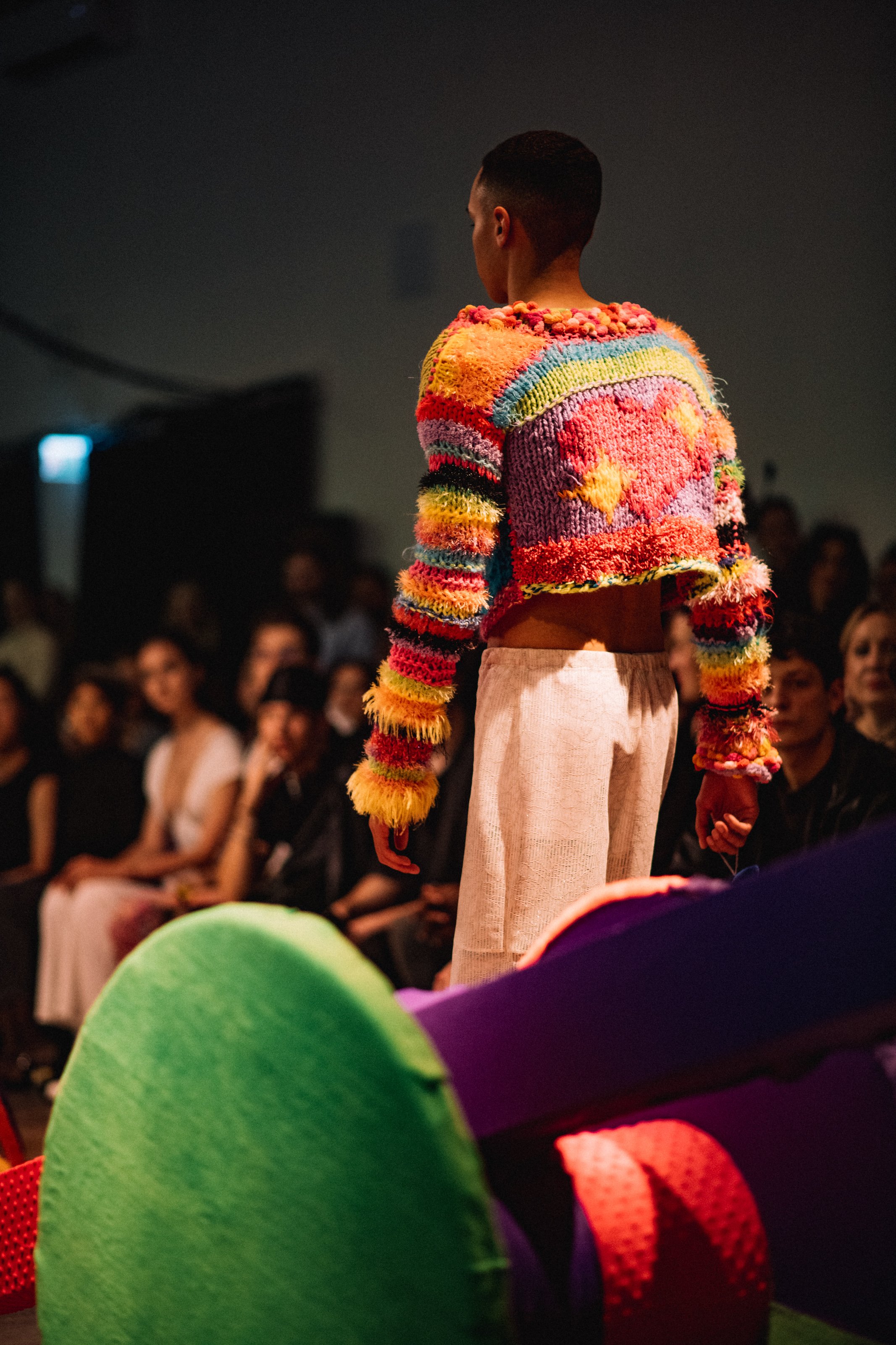 A person walking on a runway during a fashion show, wearing a colorful, textured knitted sweater with various bright shades and patterns, and loose light-colored pants.