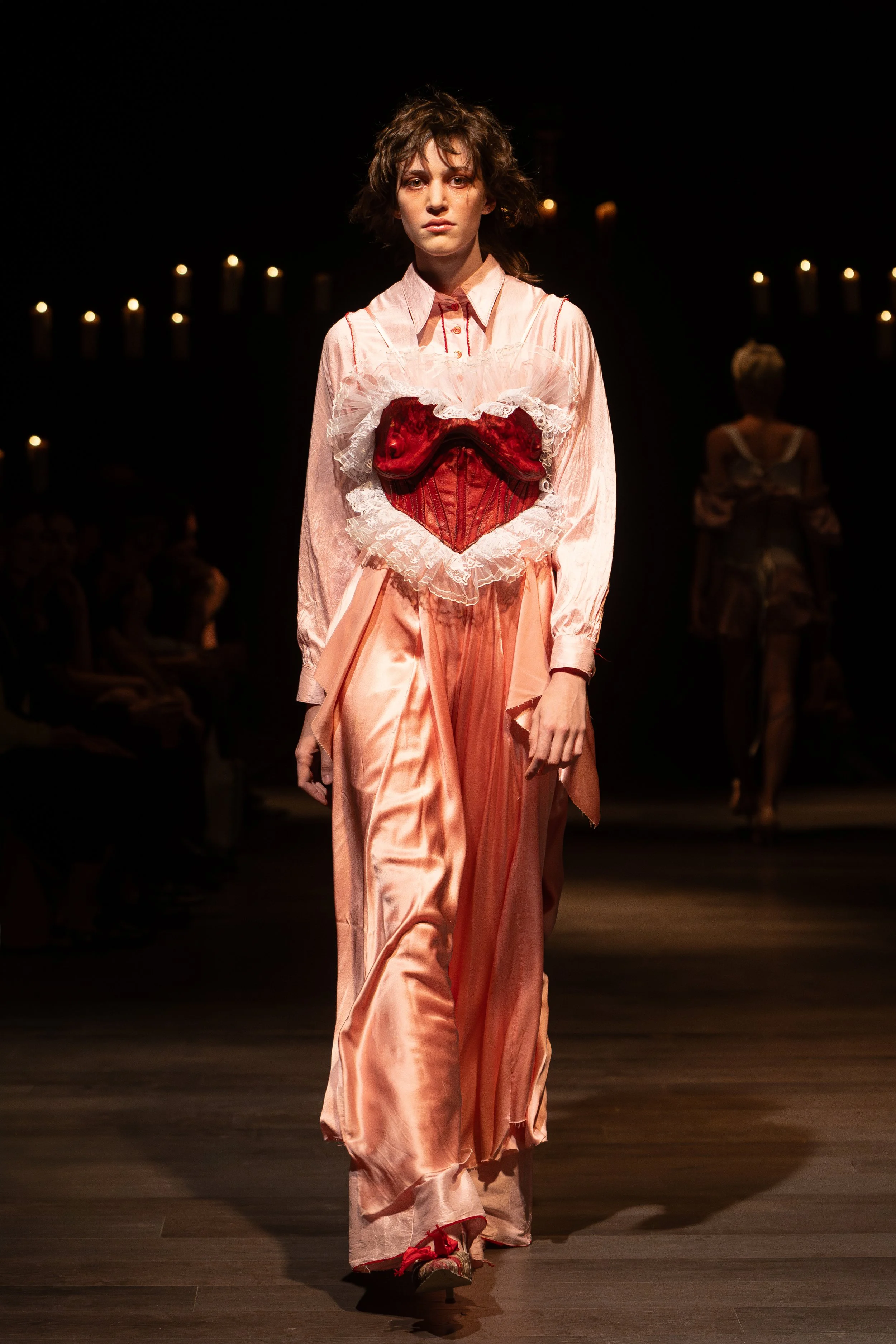 A model walking down a runway wearing a pink satin dress with red and white lace details, including a heart-shaped corset, and red shoes. The background is dark with small lit candles, and another model is visible in the distance.