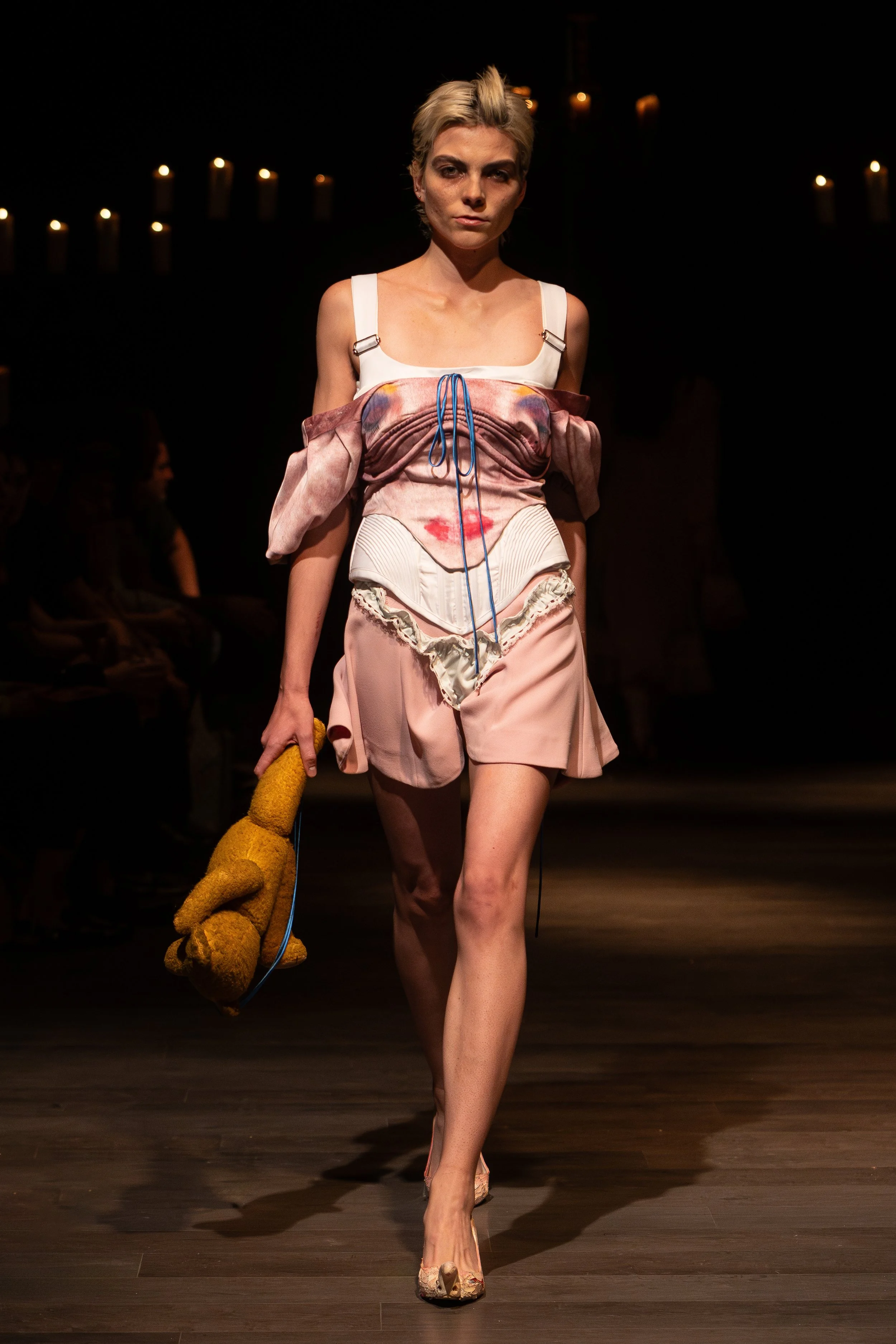 A model walks down a runway during a fashion show, wearing an avant-garde outfit with a mix of pastel colors and unique textures, holding a plush yellow stuffed animal.