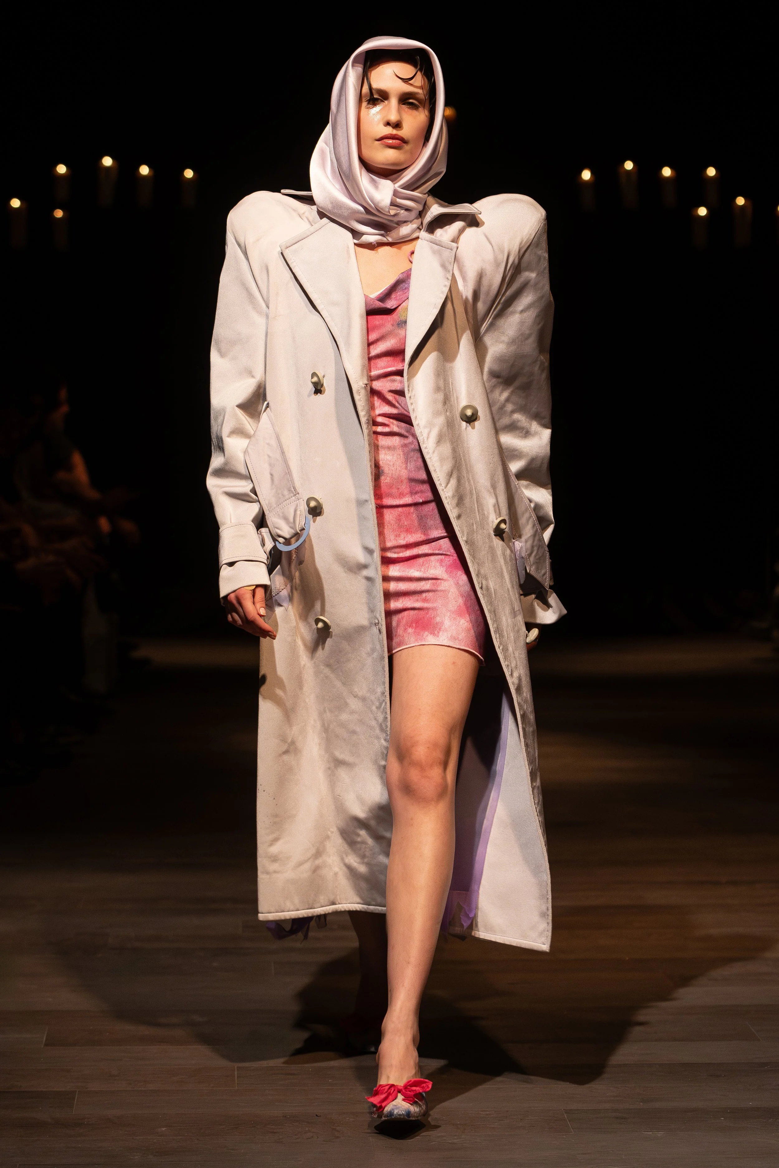 A woman walking on a runway wearing a beige coat over a pink velvet dress and red high heels with bows. She has a white scarf draped over her head and shoulders, and her makeup is minimal with bold lips. The background is dark with blurred lights and