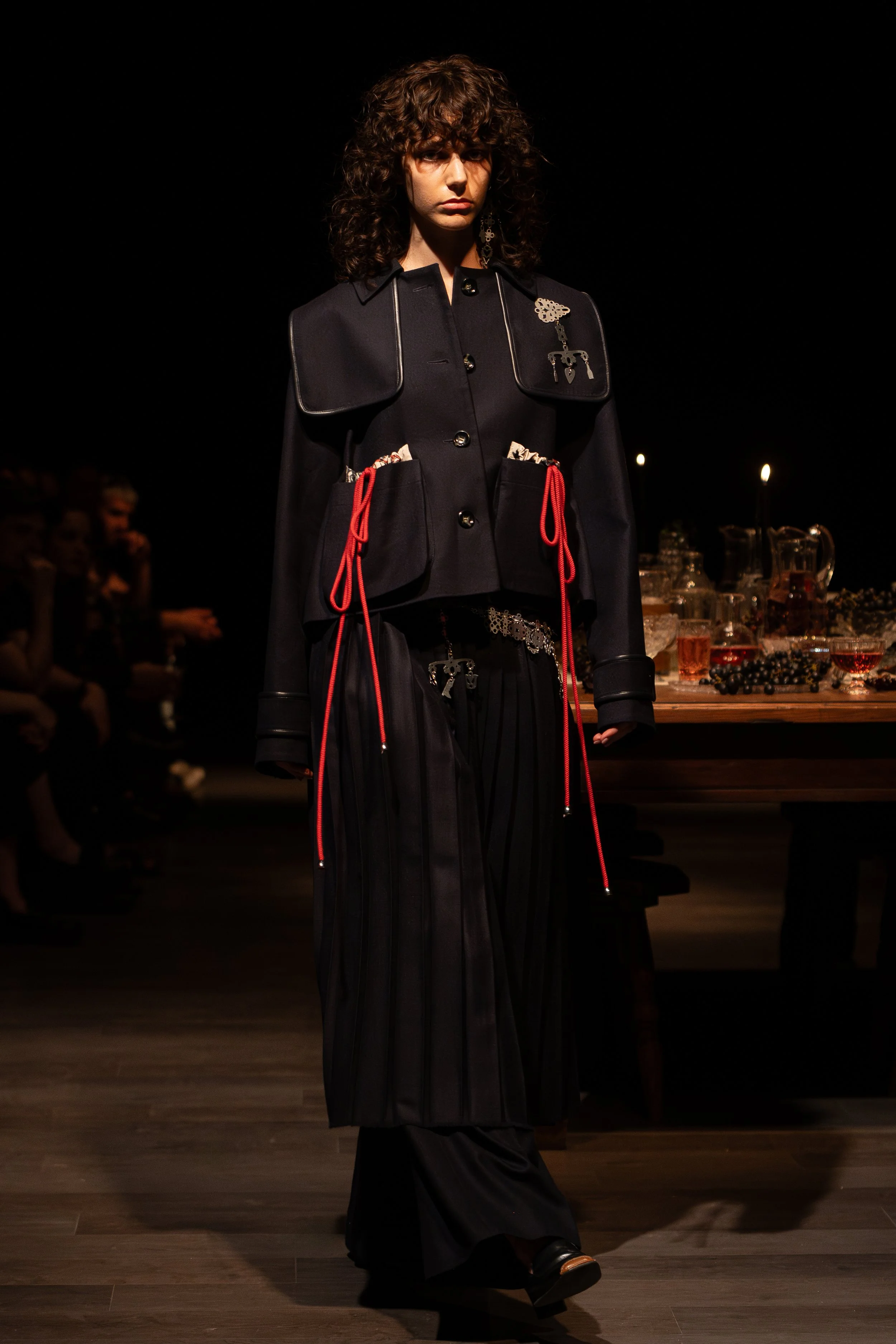 Model walking on runway during fashion show, wearing black outfit with metallic details, in a dark venue with audience watching.