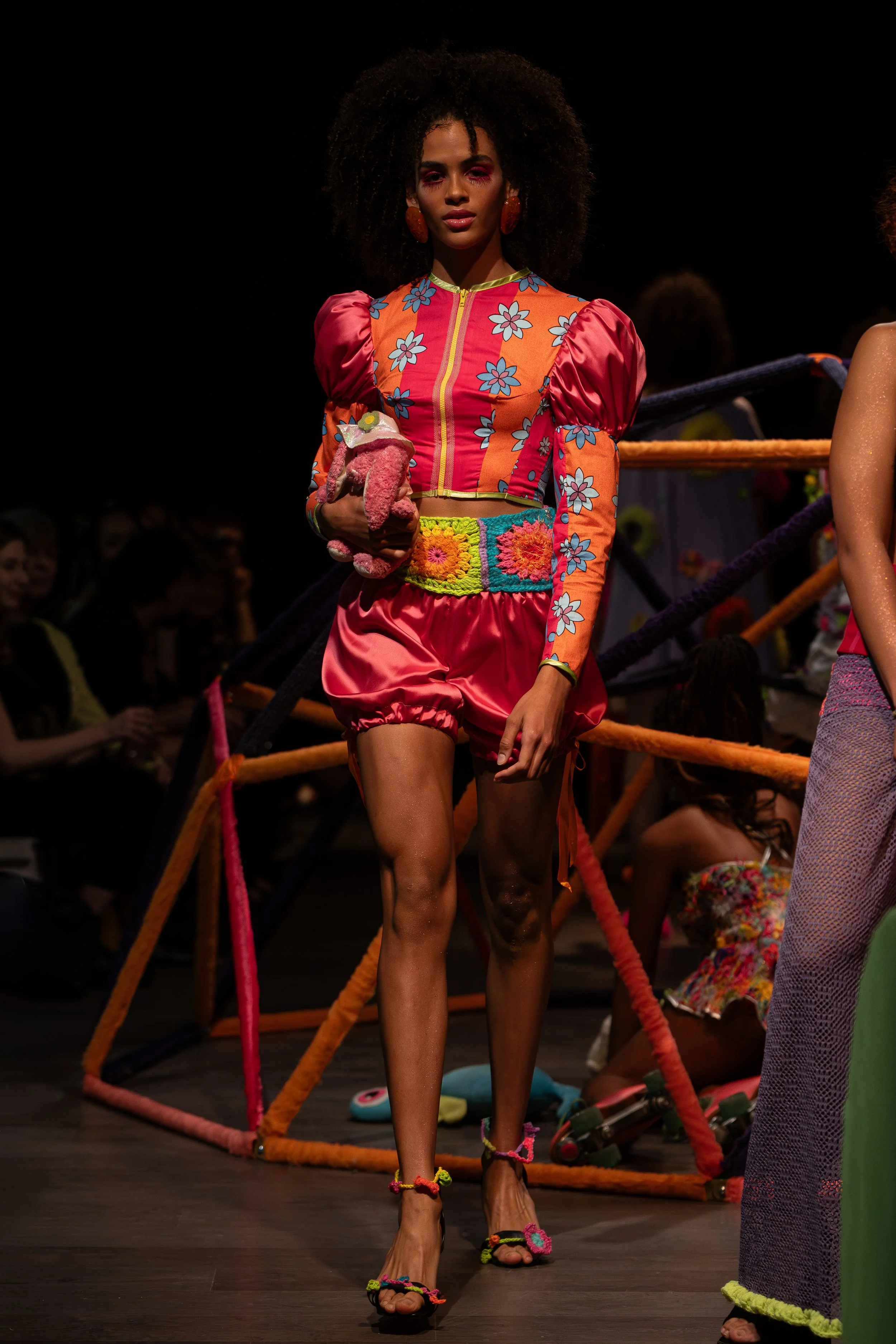 A woman with dark curly hair wearing a colorful floral outfit, holding a small pink stuffed animal, walking on a runway at a fashion show.