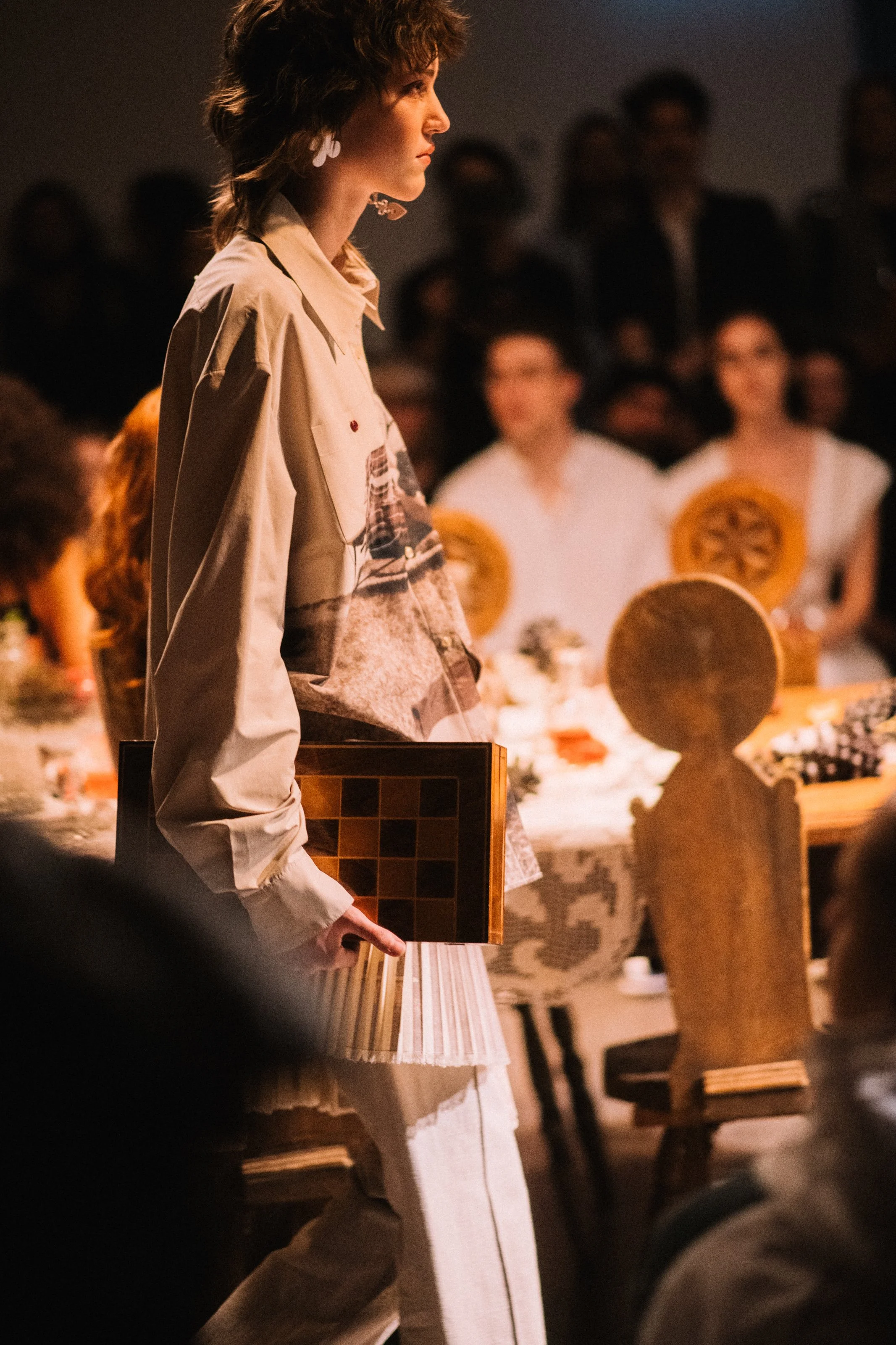 A woman with short brown hair wearing a beige jacket and a print shirt is standing in profile at a table during an event, holding a wooden board. There is a blurred crowd in the background in a dimly lit environment.