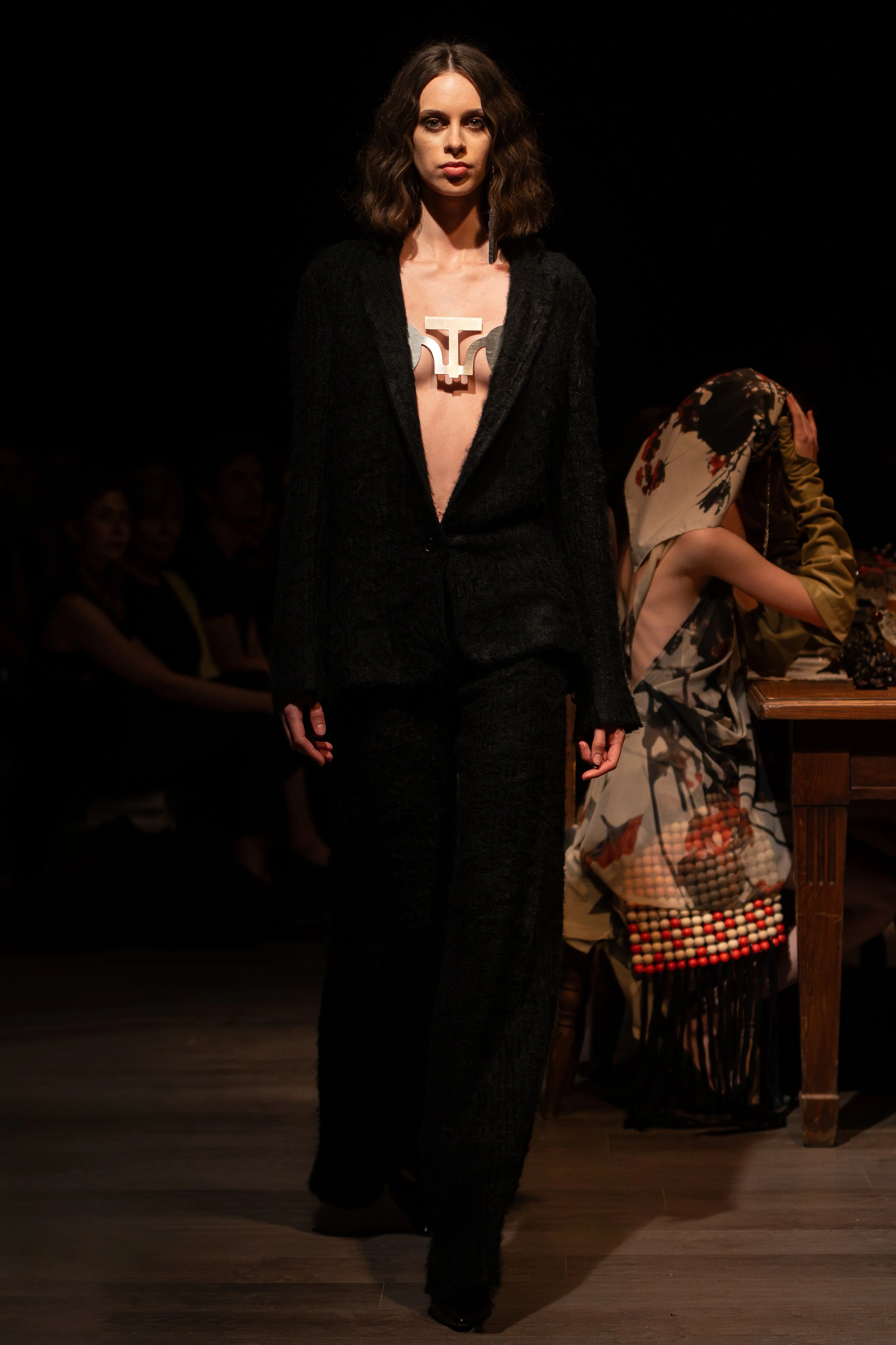 A woman walking on a runway during a fashion show, wearing black pants and jacket with a large geometric necklace, with a seated audience in the background.