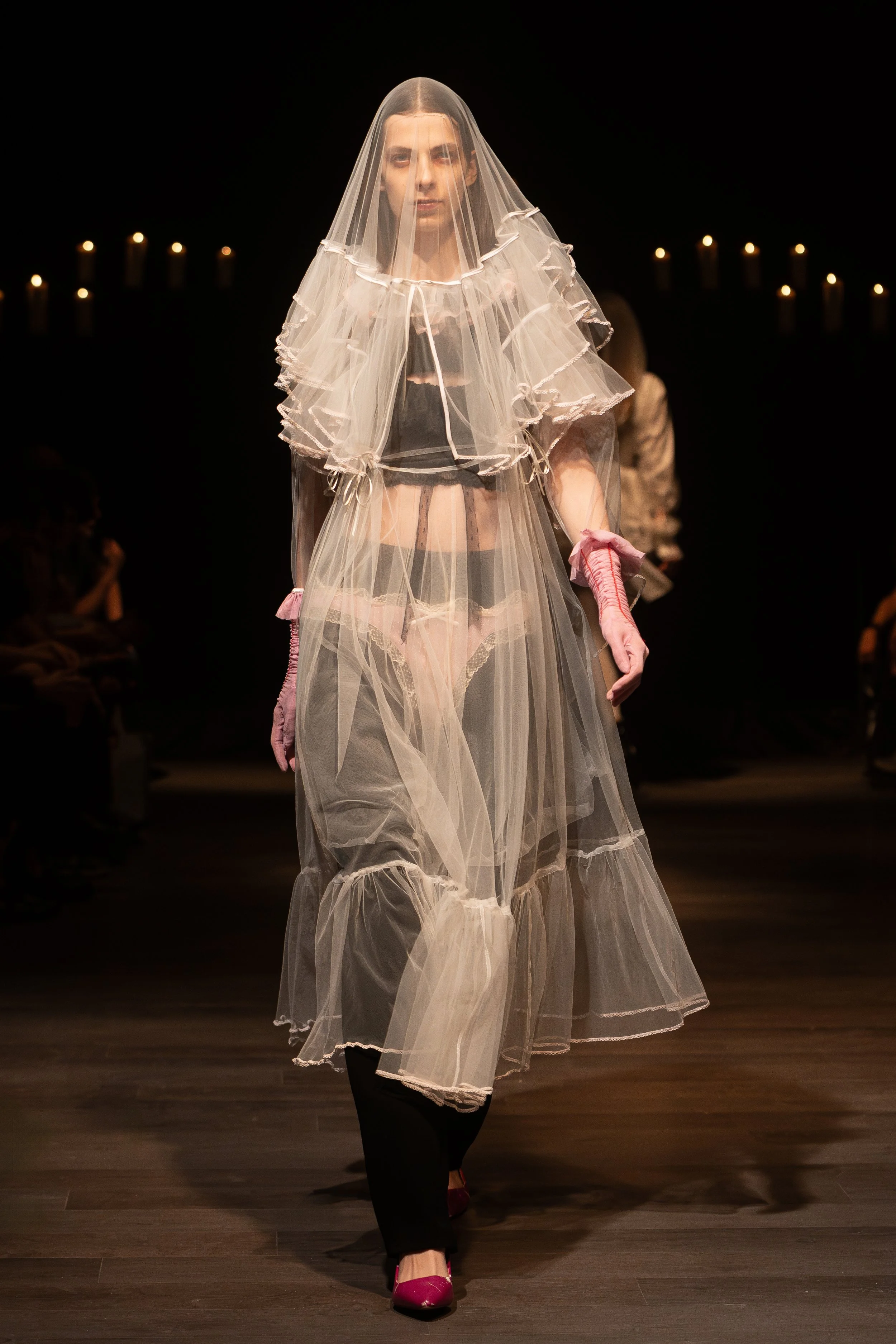 Model walking on runway wearing sheer white layered dress with ruffled edges, black lingerie underneath, pink gloves, and pink high heels, with candles in the background.