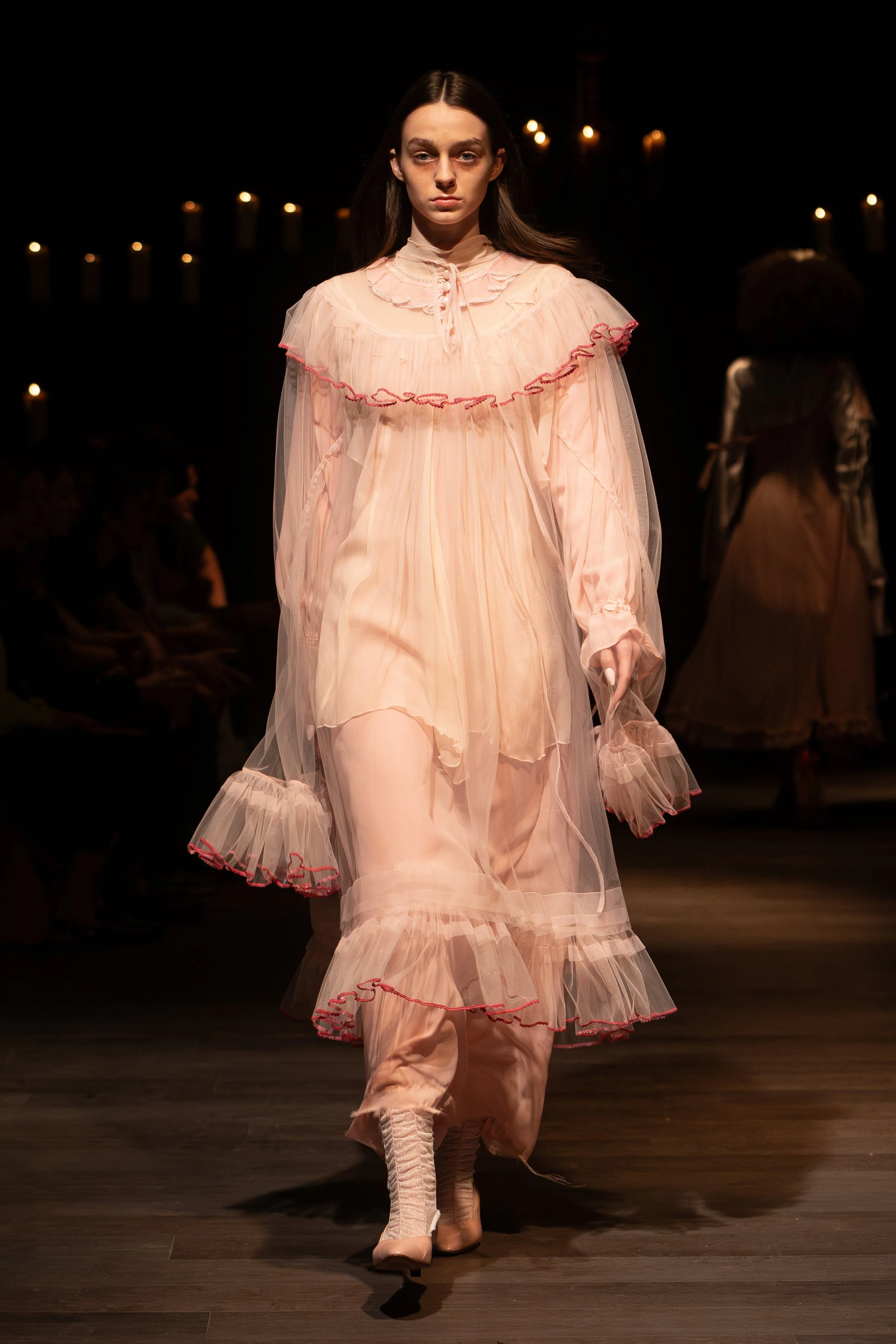 A female runway model wearing a light pink, ruffled, sheer dress with matching high heels, walking on a wooden runway during a fashion show under dim lighting with candles in the background.
