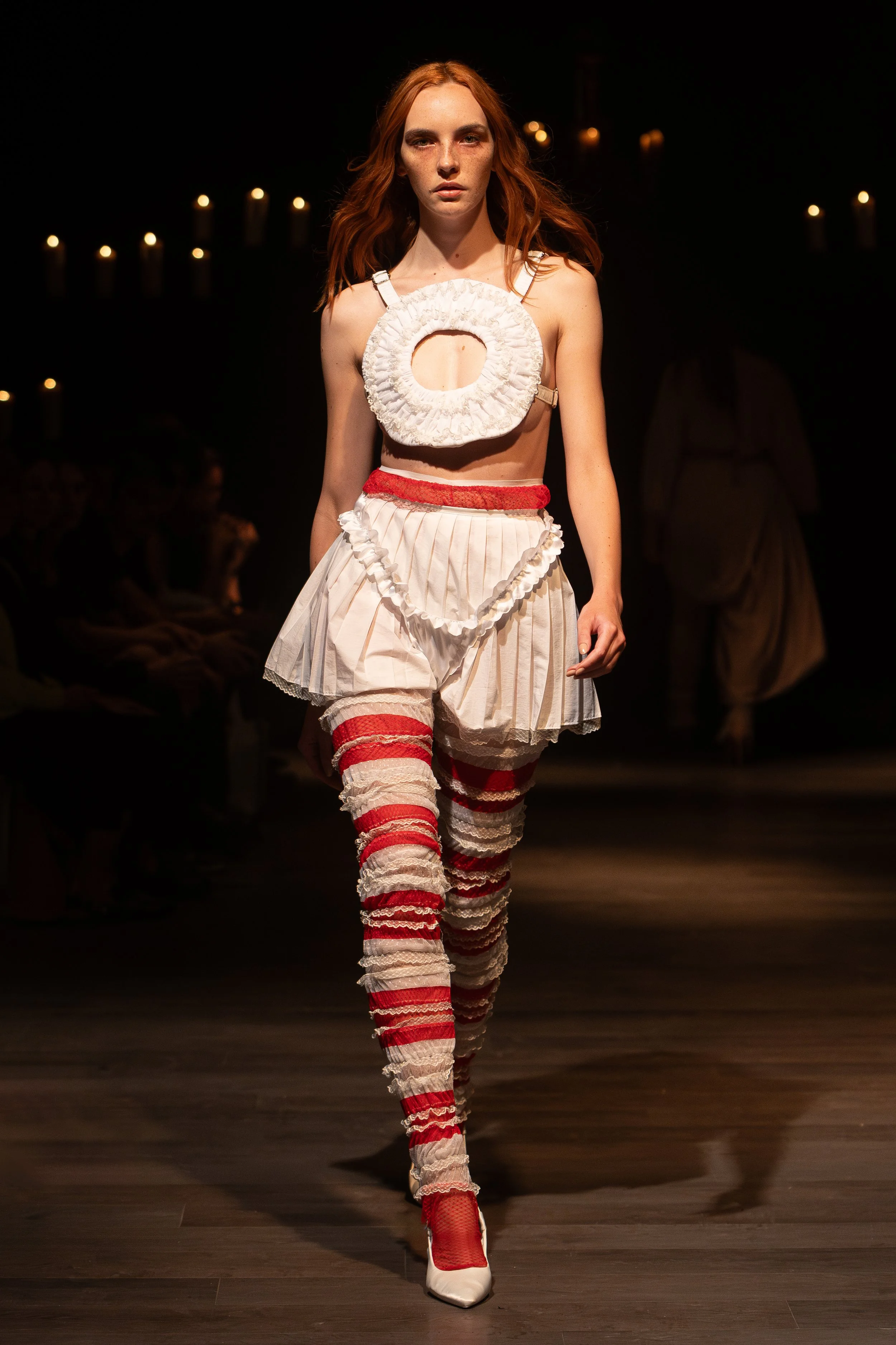 A red-haired female model walking on a runway with a dark background and string lights. She is wearing a unique white dress with red and white striped thigh-high socks, white shoes, and a circular white ruffled accessory on her chest.