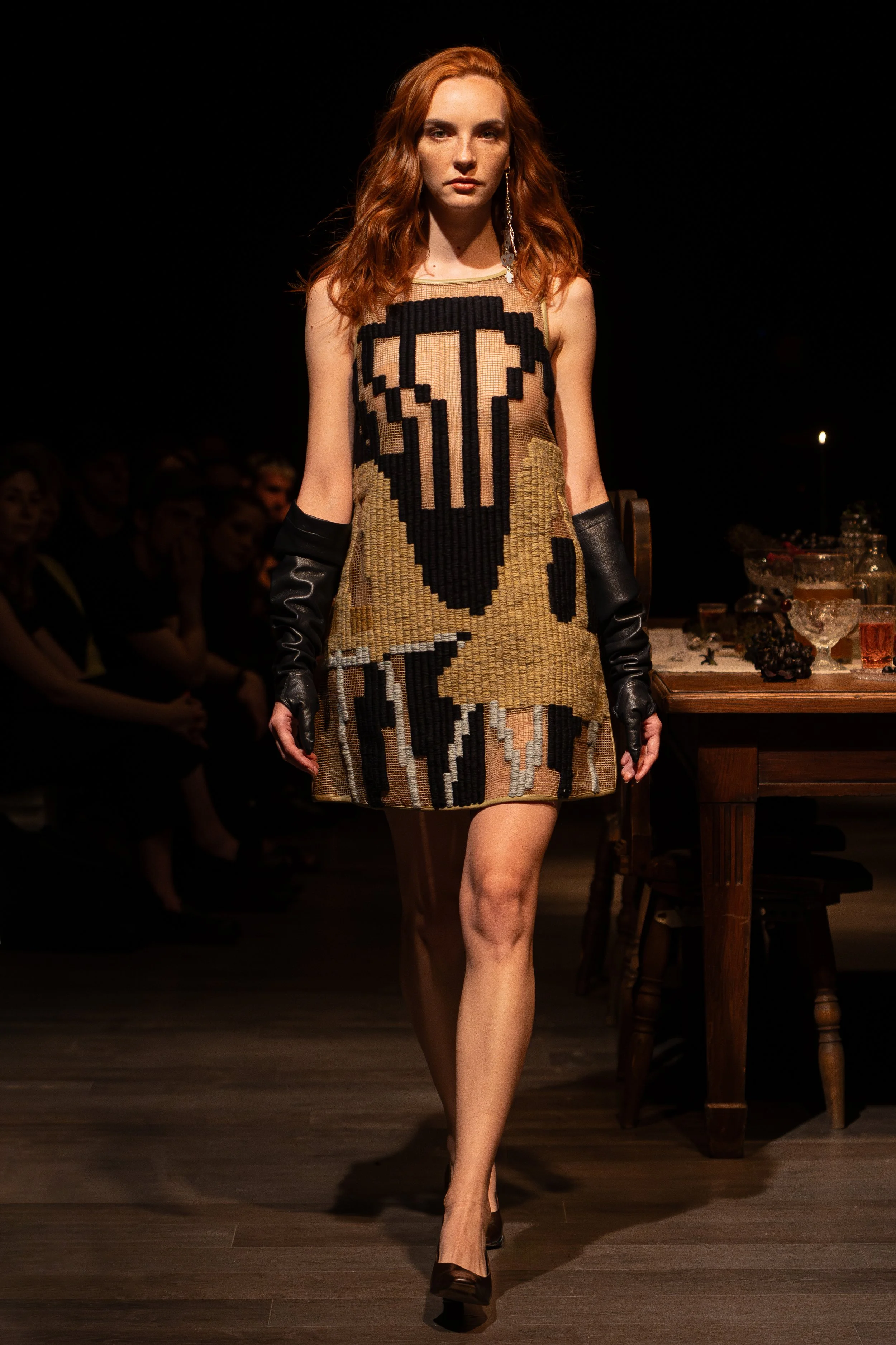 A woman with red hair walking on a runway wearing a dress with pixel art skull design and long black gloves.