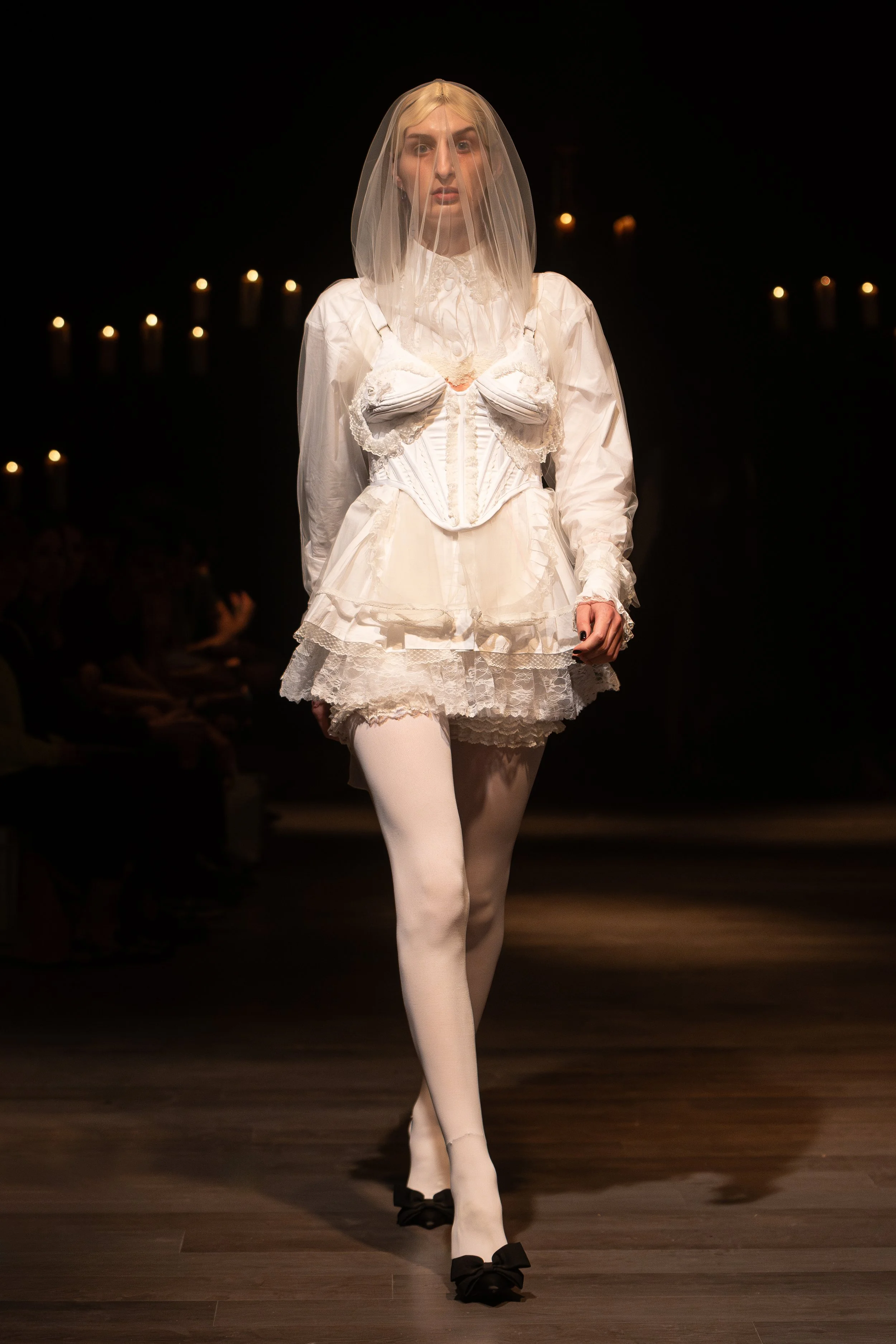 A female model walks down a runway in an all-white vintage-inspired bridal gown with a lace-trimmed skirt, corset-style bodice, and a sheer veil covering her face, with dark background and candles in the distance.