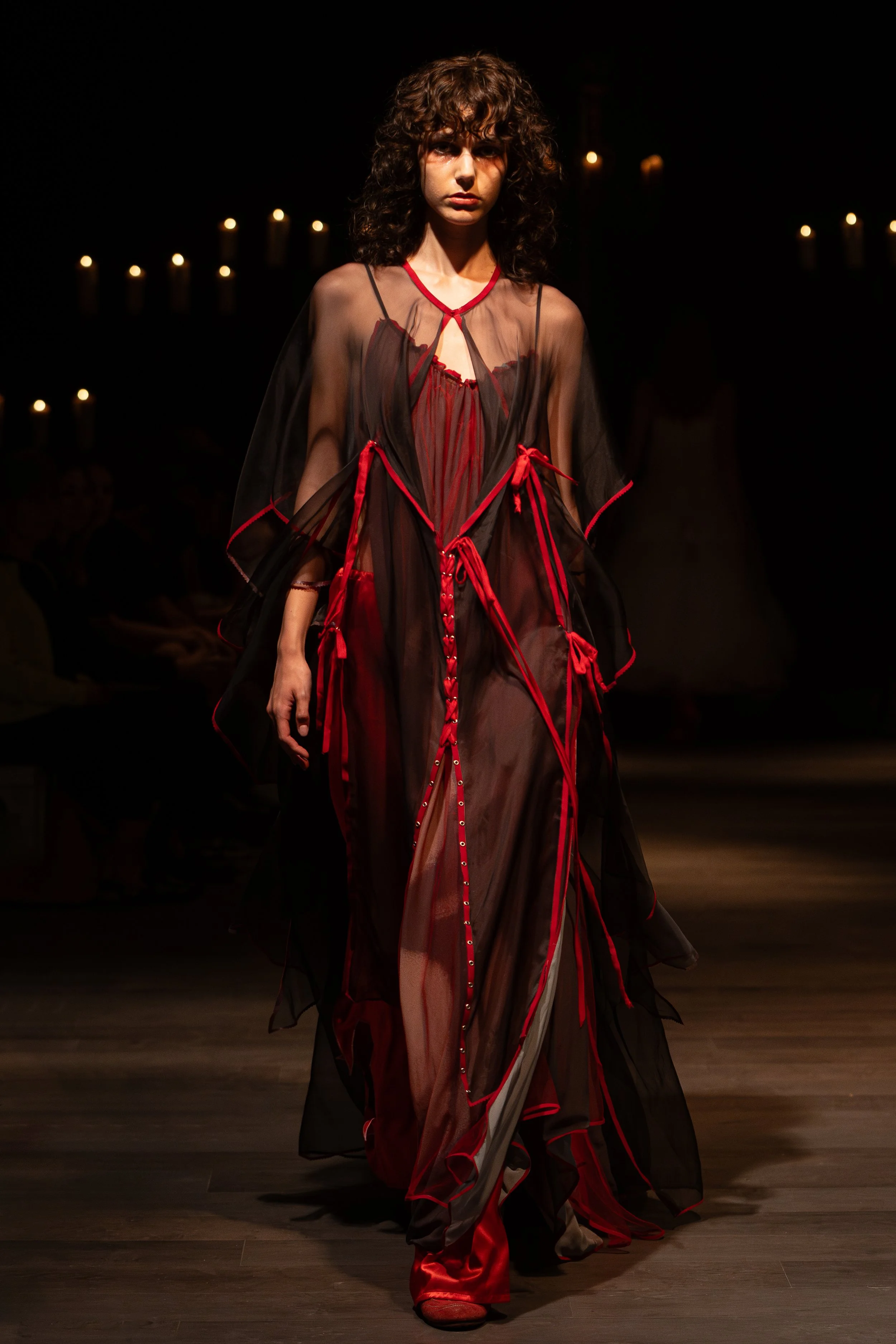 Model walking on runway in sheer black and red gown with red ribbons, lighting focused on her, dark background with candles.