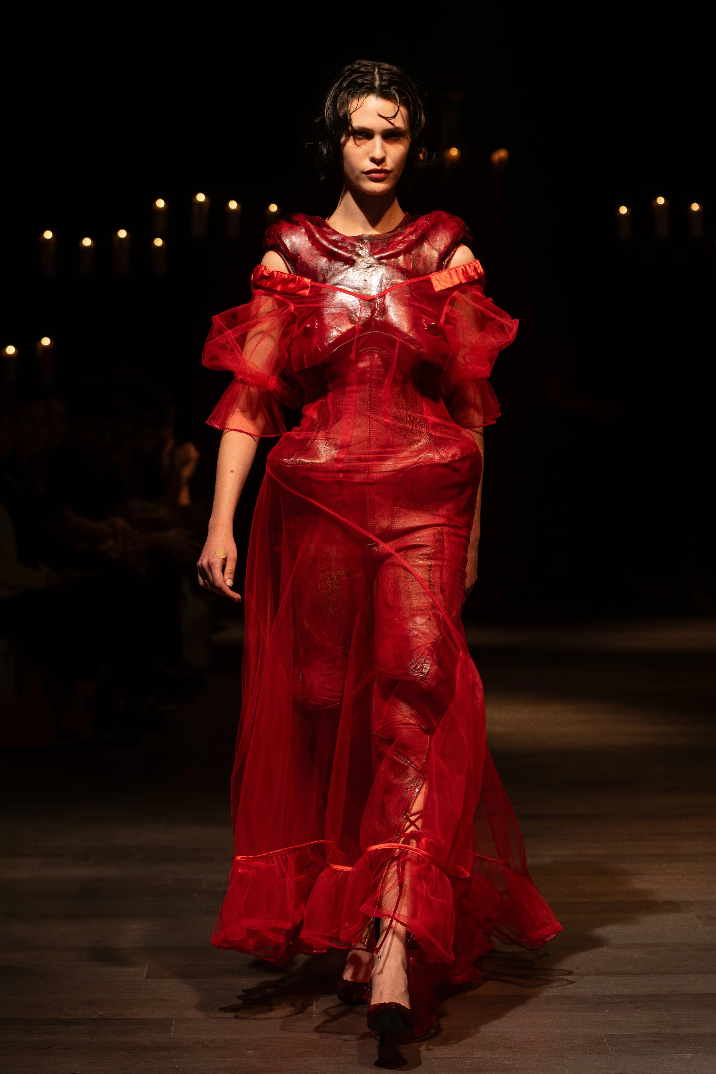 A female model walks down the runway wearing a flowing red dress with sheer tulle and leather accents, dark hair, and dark eye makeup, illuminated by a spotlight in a dark setting with candles in the background.