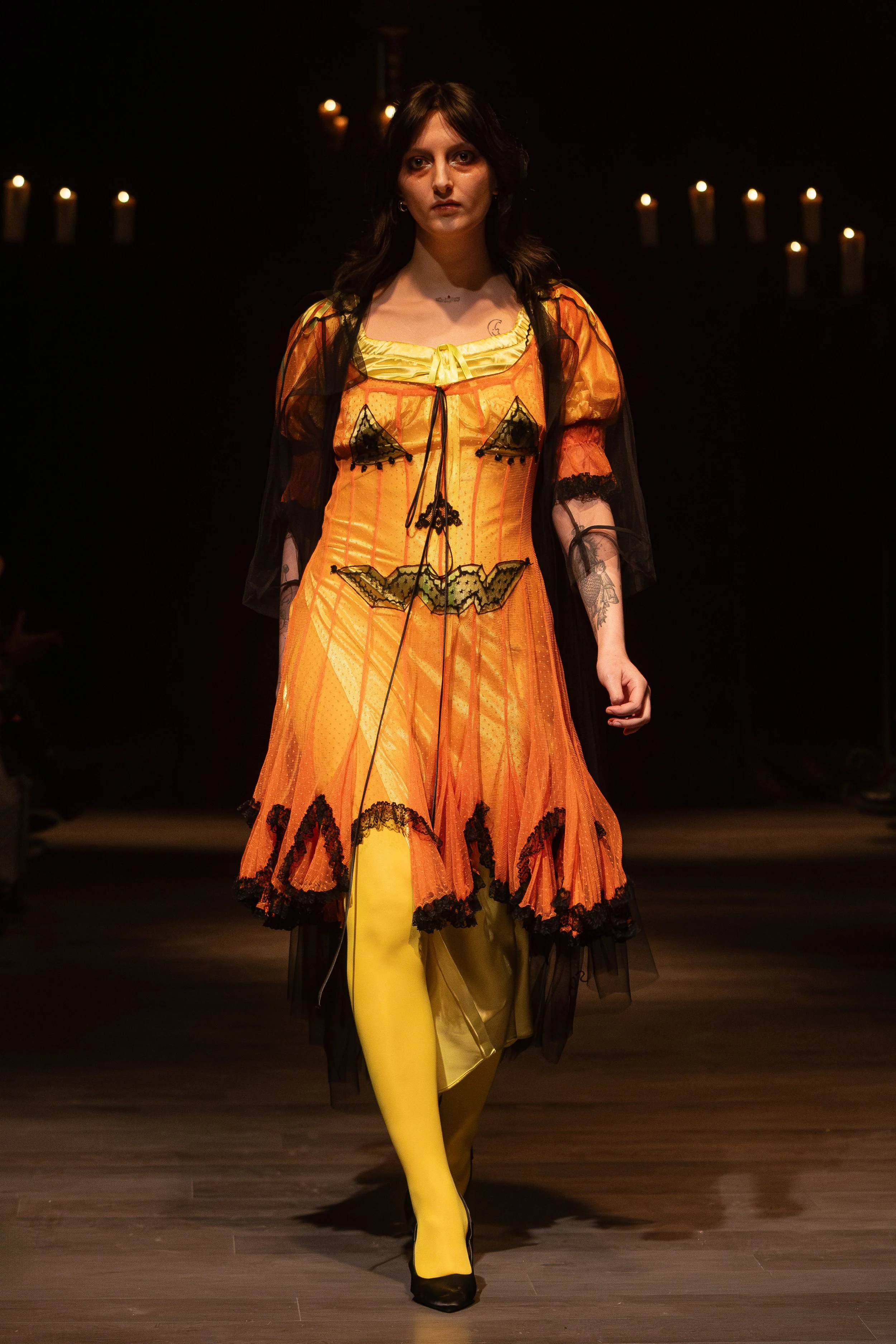 A woman walking down a runway in a colorful, Halloween-themed dress with orange, black, and yellow details, yellow tights, and black shoes. The background is dark with candles.