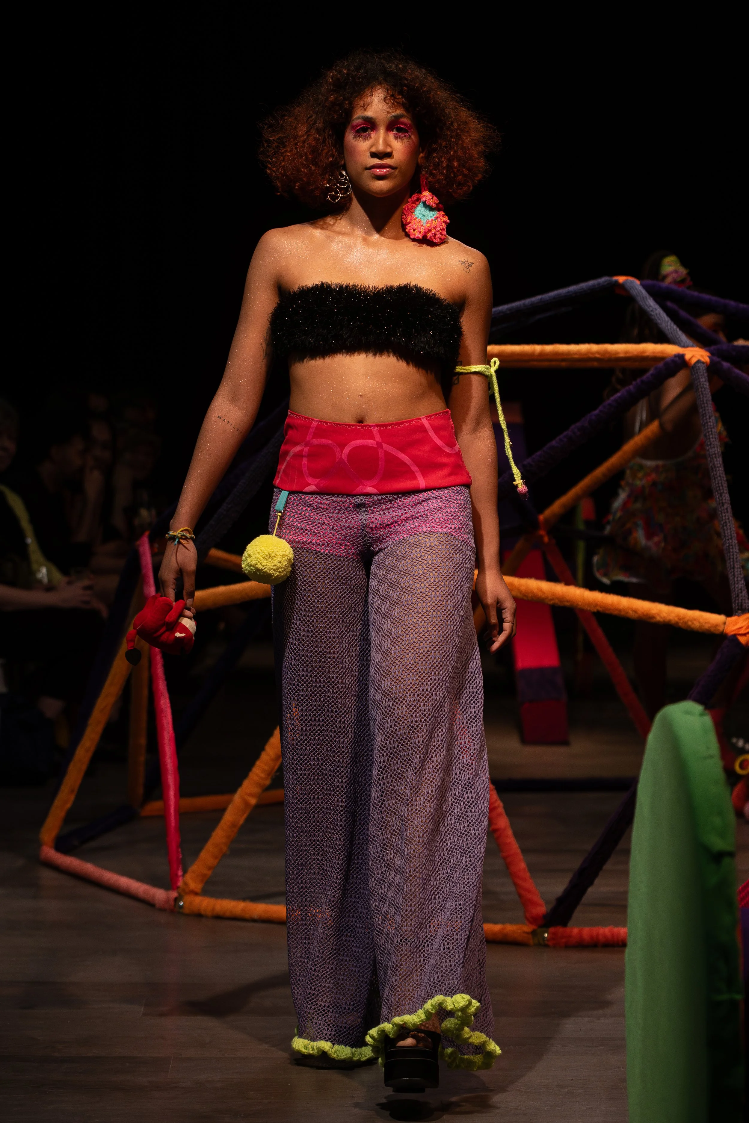Model walking on runway at fashion show, wearing colorful outfit with fishnet pants, fluffy black top, and flower earrings; bright makeup, dark background, audience in dim lighting.