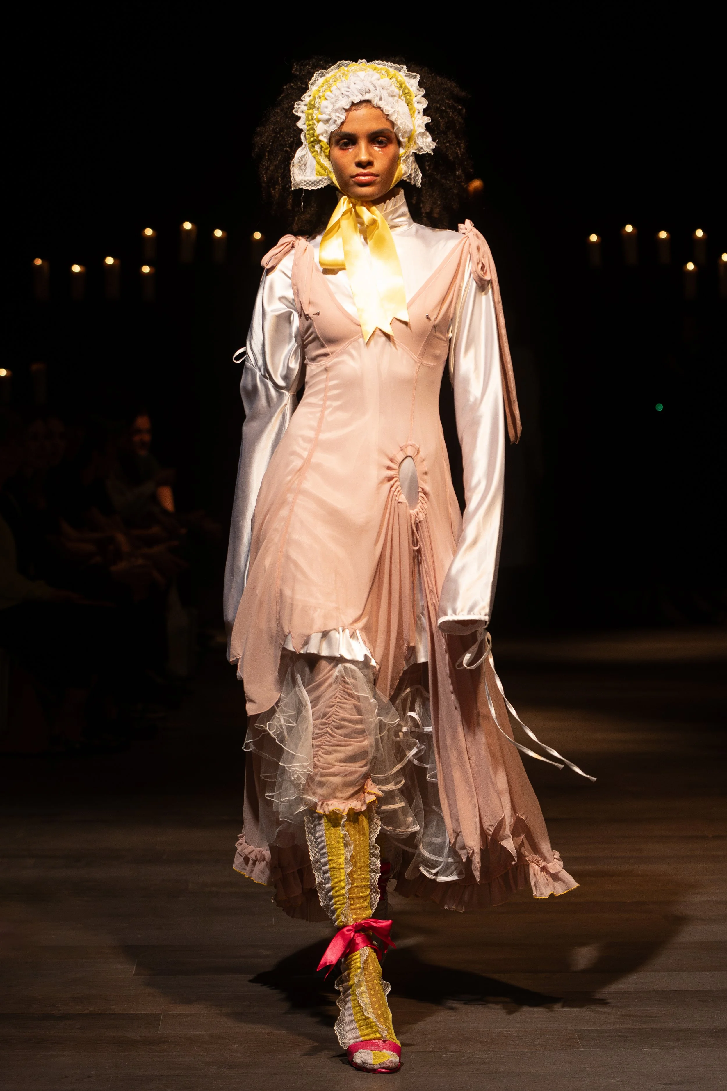 Model walking down runway in vintage-inspired pink and cream dress with lace, ruffles, and bows, wearing colorful socks and sneakers, with dark background and audience in the shadows.