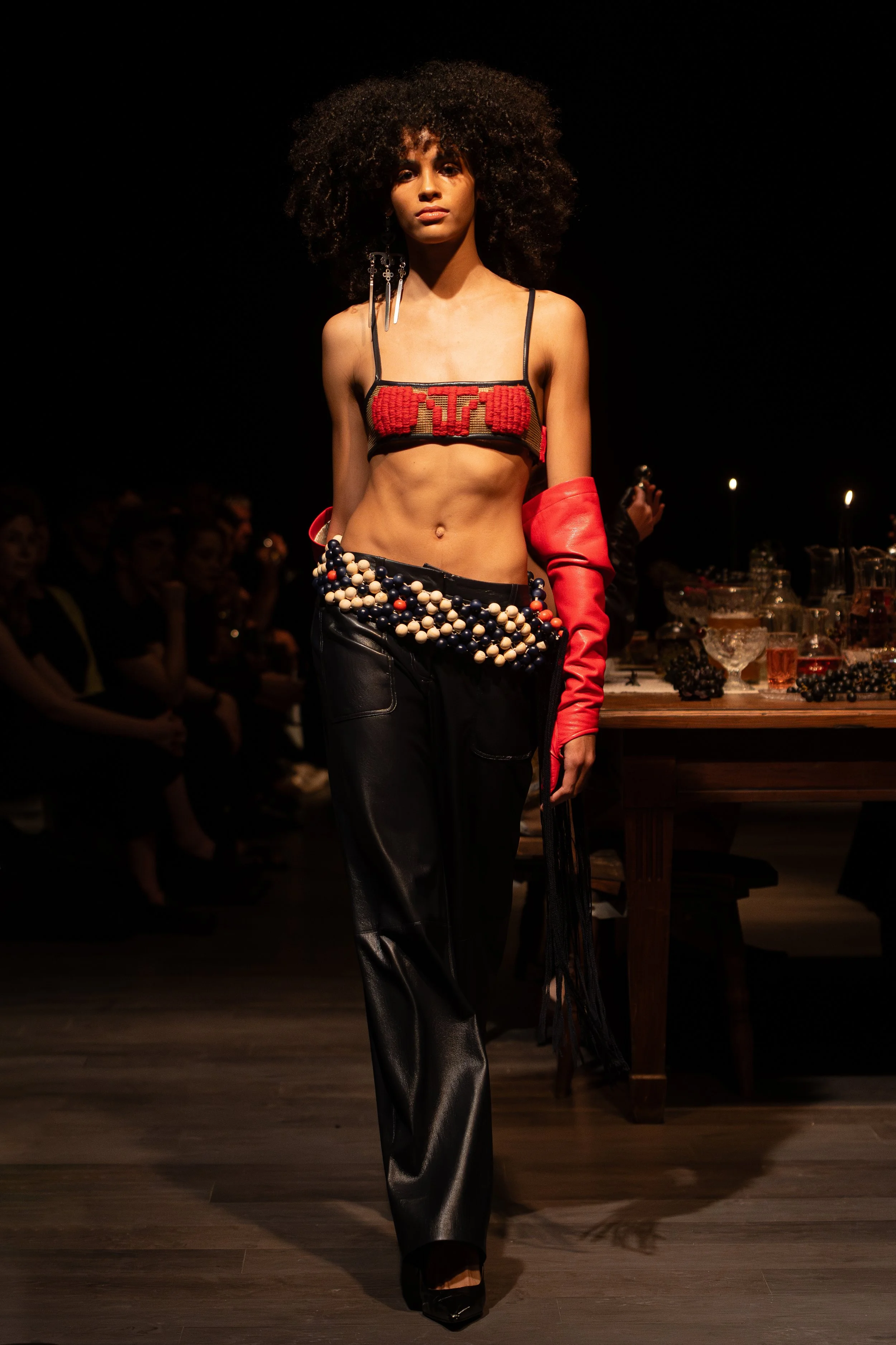 A female model walking on a runway during a fashion show, wearing black leather pants, a decorated crop top with red and gold embroidery, a red arm-length glove on her left arm, and black high heels. Audience members are visible in the background.
