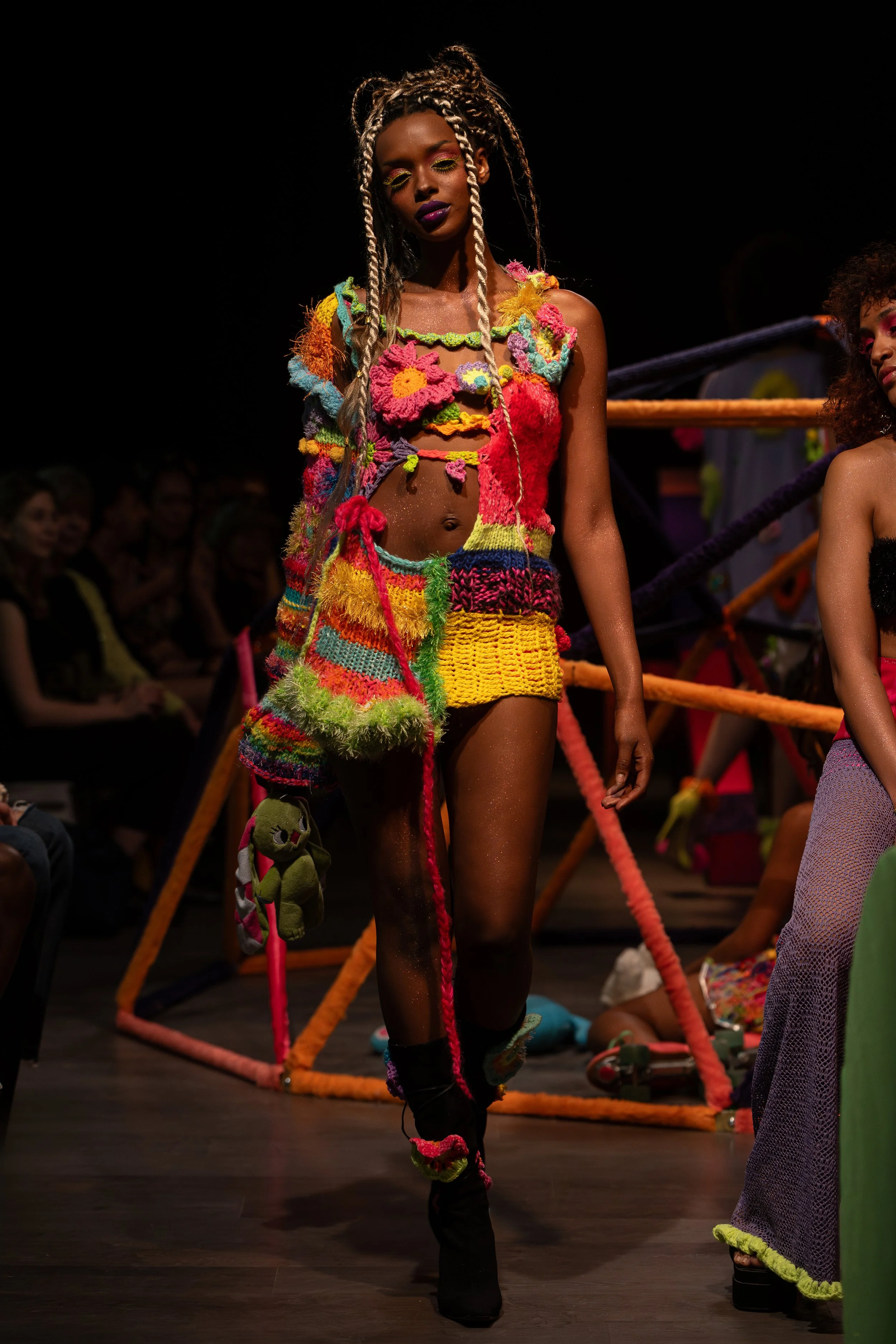 A woman with dark skin and blonde braided hair walking on a runway, dressed in colorful, textured, crochet-style clothing and accessories, with a black background and spectators in the audience.