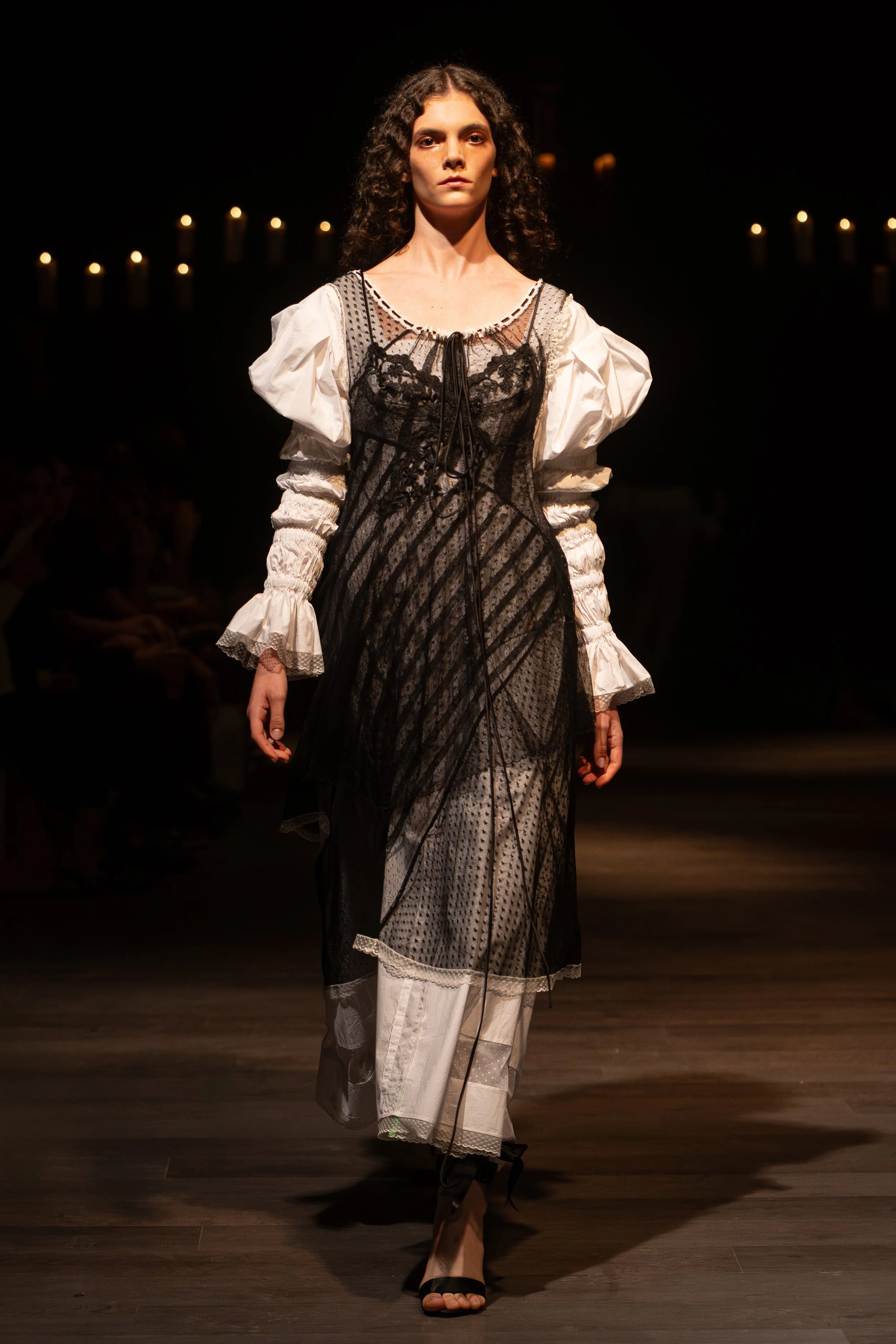 A female model walking down a runway during a fashion show, wearing a layered outfit with a black lace dress over a sheer top, paired with white pants with lace details at the hem, and black high heels. The background is dark with dim lighting and ca