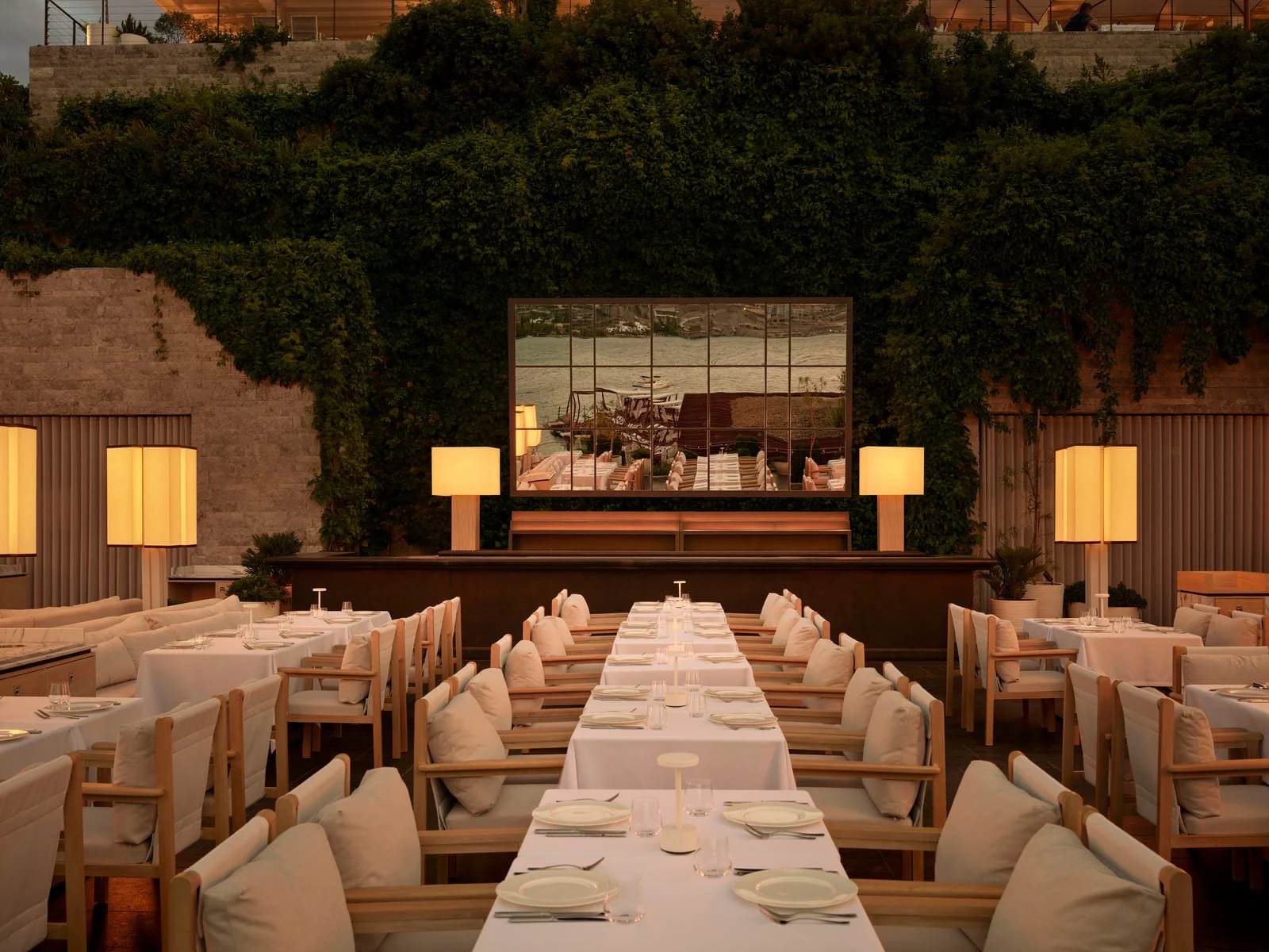 Elegant restaurant dining area with white tablecloths, cushioned chairs, and large beige lamps, featuring a large mirror on a green wall reflecting outdoor furniture and water.