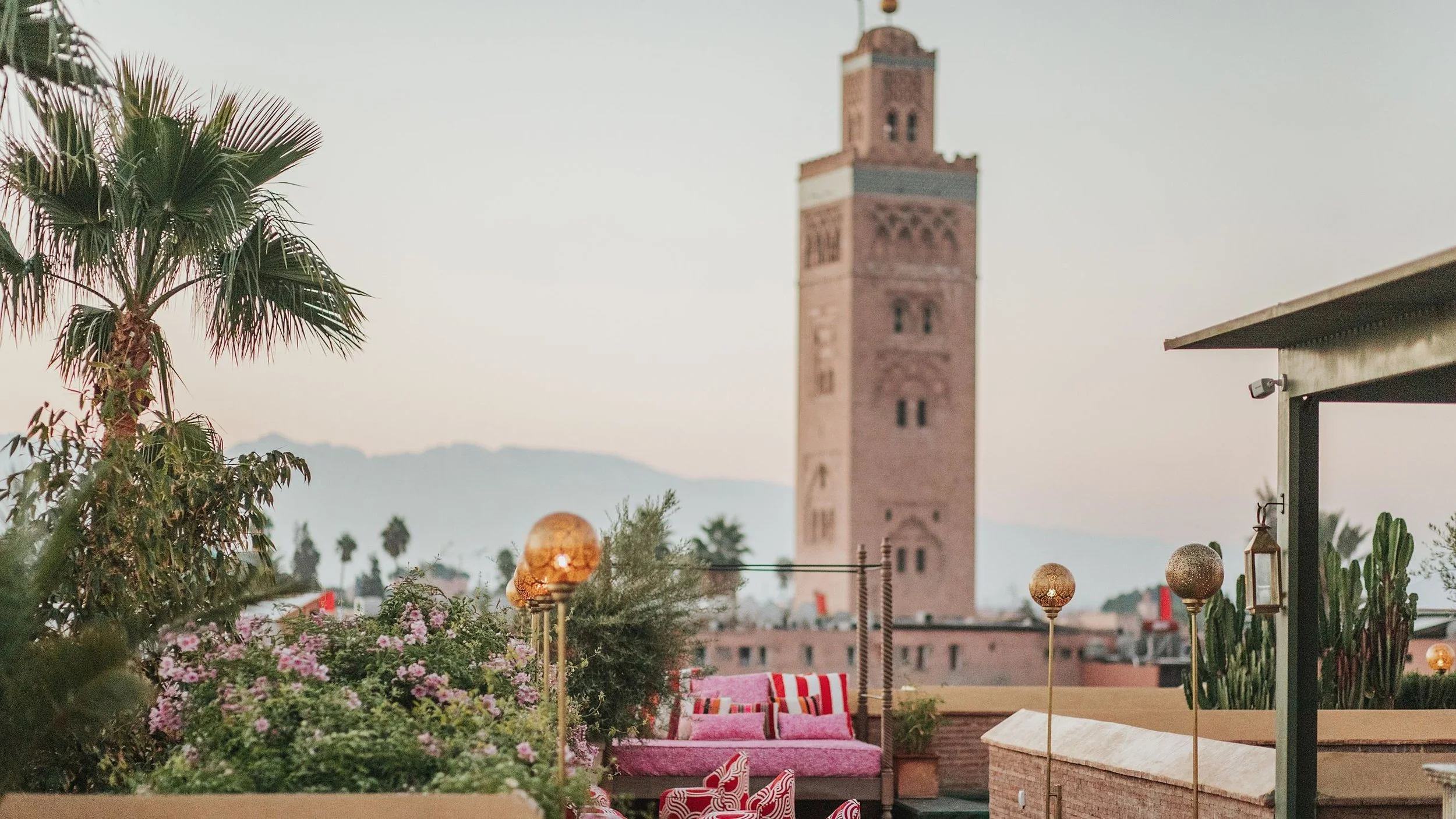 Rooftop terrace with pink seating, gold lamps, lush plants, and a view of a tall brick tower in the distance at sunset.