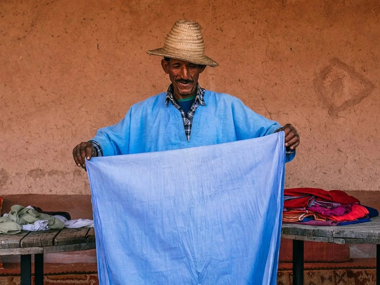 An elderly man wearing a straw hat and a blue shirt holding a large piece of light blue fabric, standing in front of a tan wall with tables of colorful fabrics.
