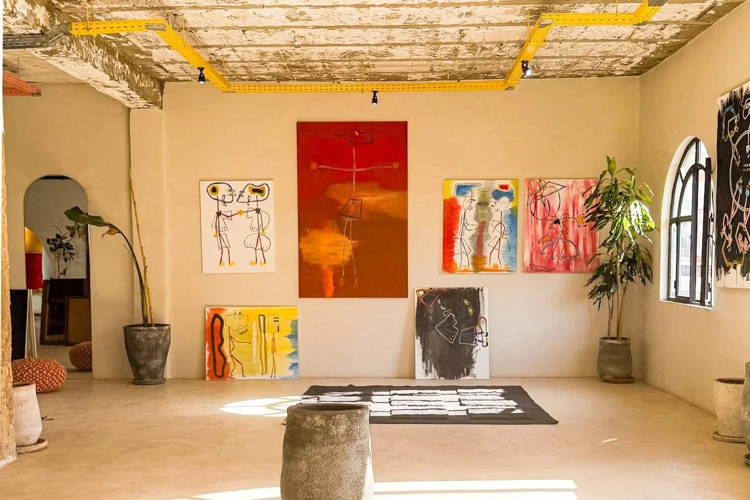 Art gallery with colorful paintings on a white wall, potted plants, and sunlight streaming through windows.