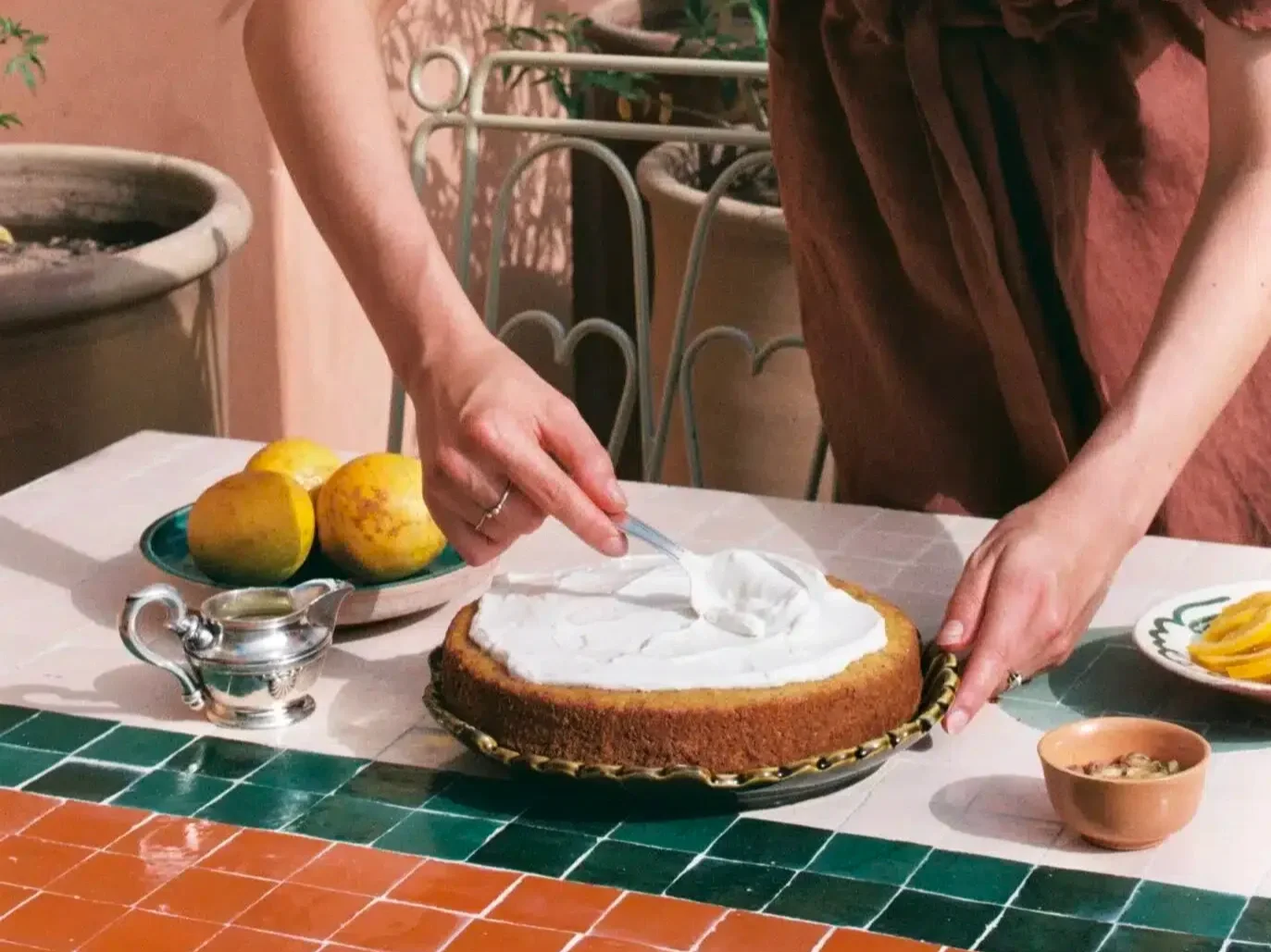 Person decorating a cake with white frosting on a plate, with lemons, a small teapot, a bowl, and a plate of sliced yellow fruit on a tiled table.