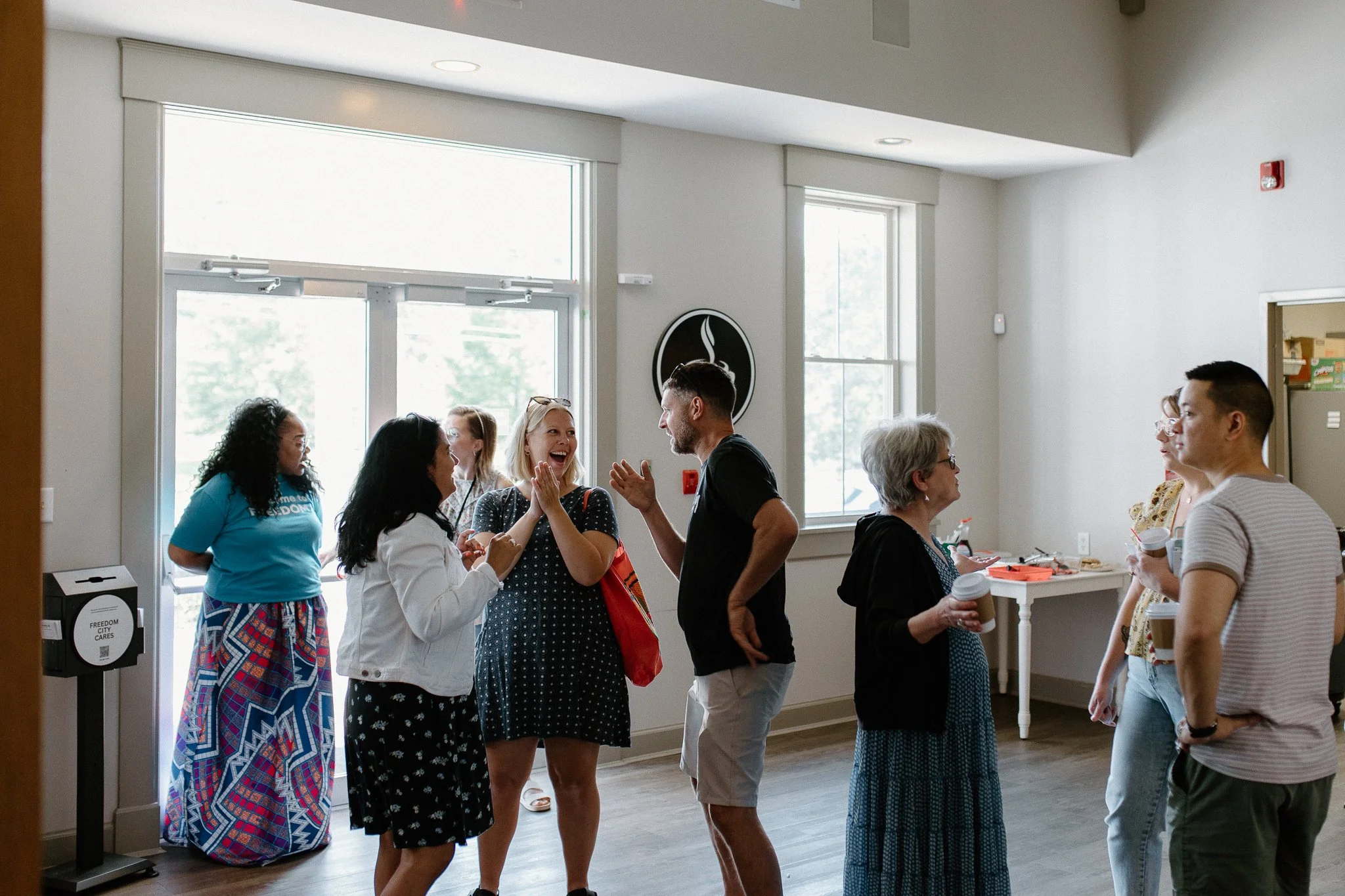 Group of diverse people chatting and smiling indoors with large windows and a coffee station.