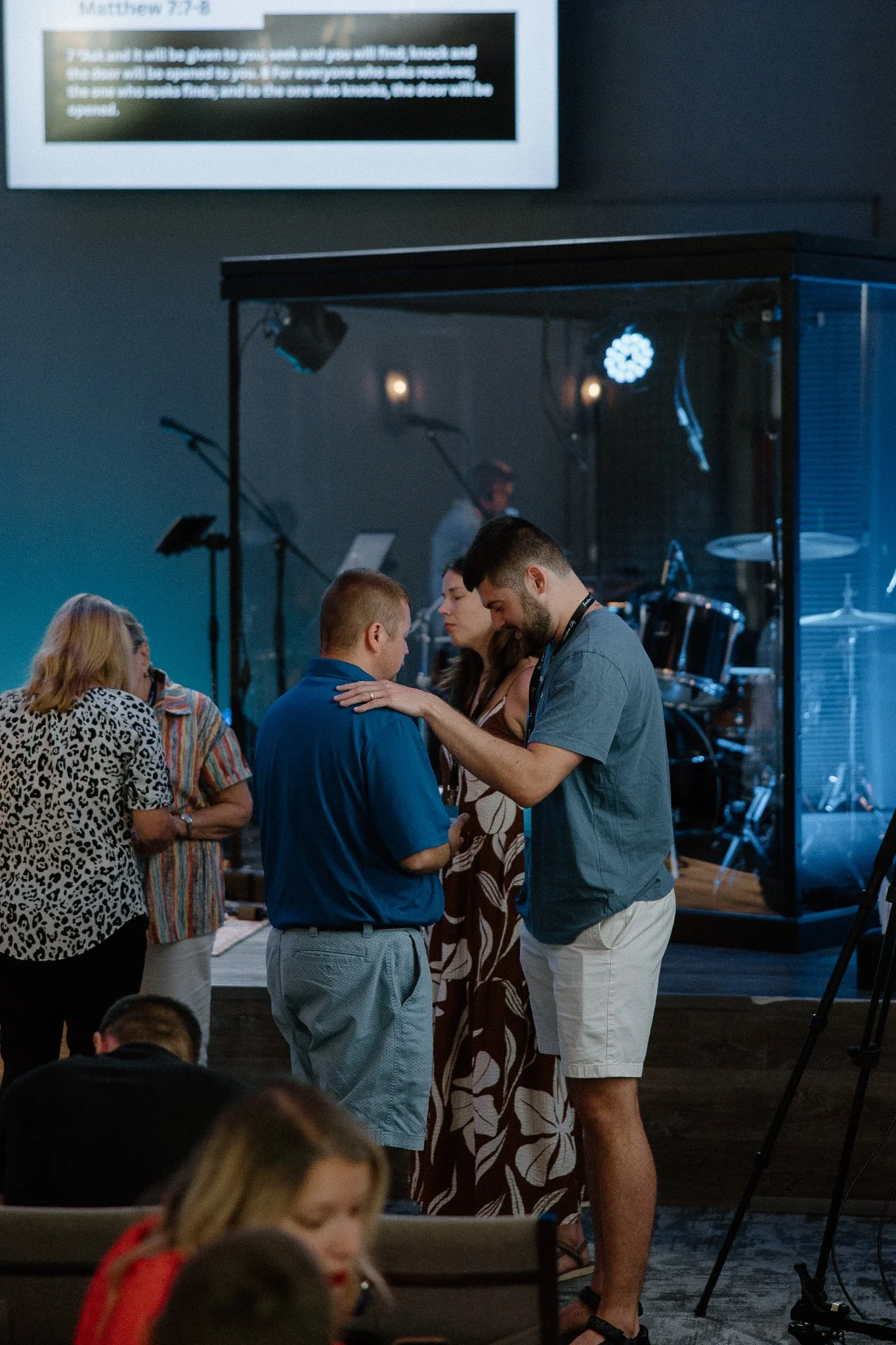 People praying or in a moment of prayer at an indoor religious event with a stage and musical instruments in the background.