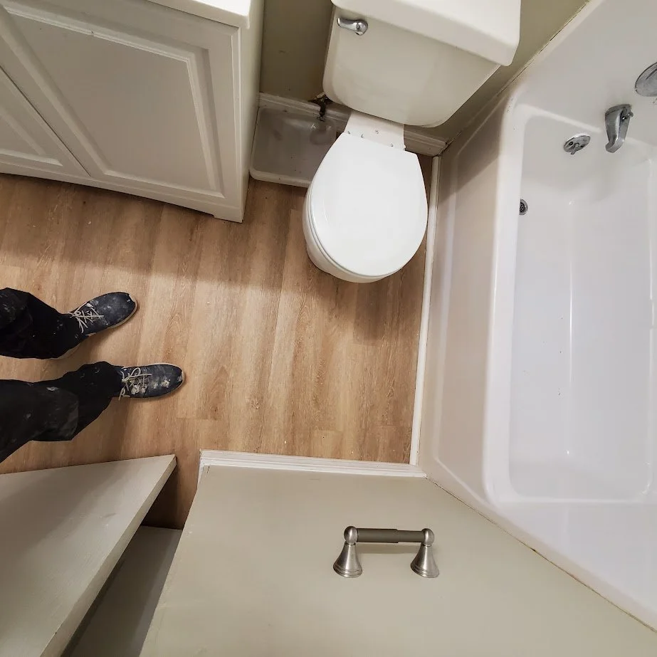 View of a small bathroom with toilet, bathtub, and wooden floor. Part of a person’s legs wearing black pants and boots are visible.