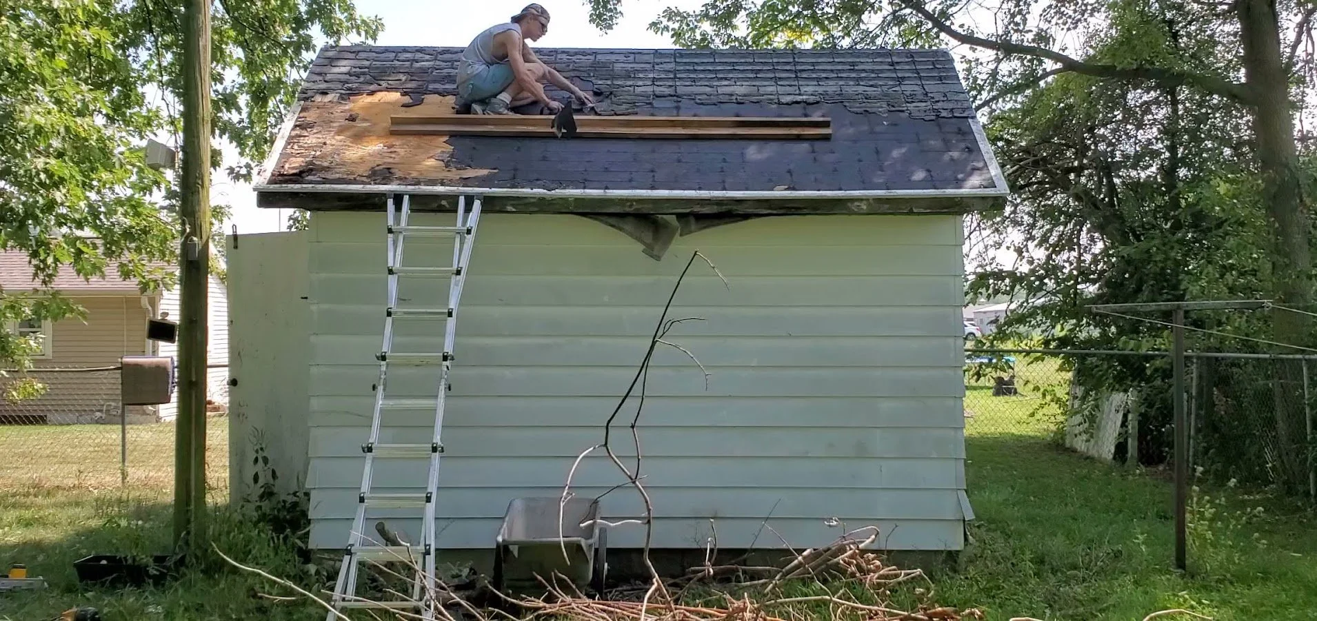 A person is working on repairing or replacing the roof of a small house, sitting on the roof with tools. The house has light-colored siding and the roof is partially stripped, with some old shingles removed, and new roofing materials nearby.