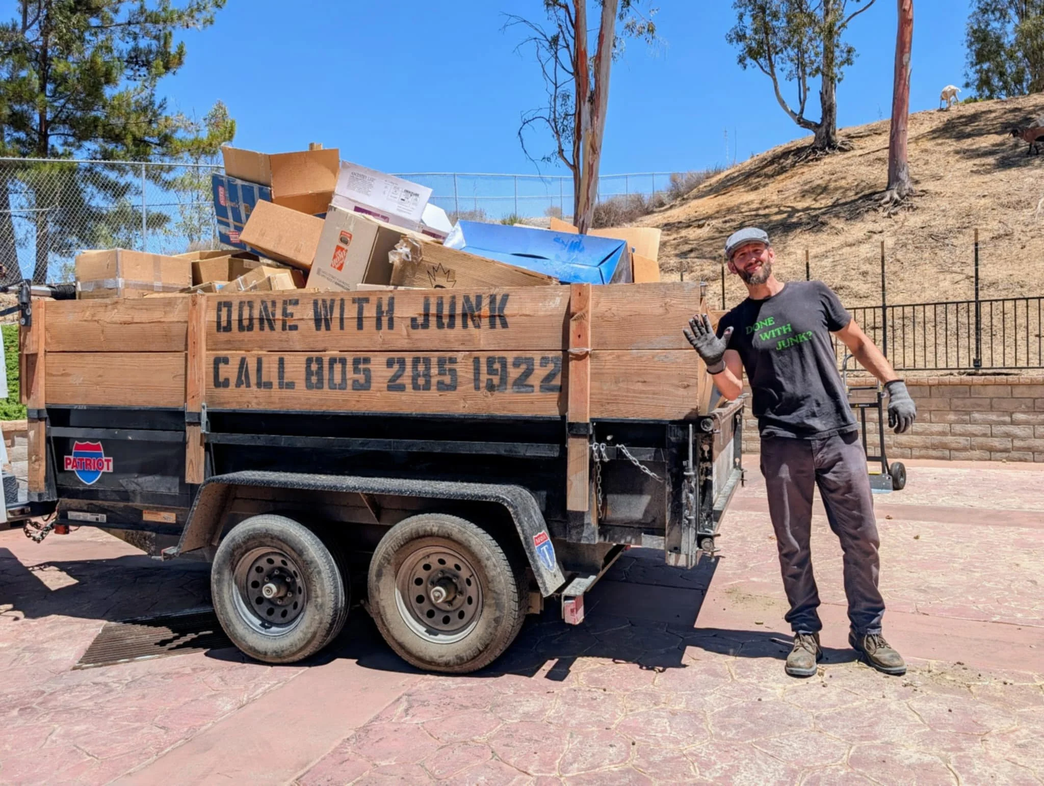 A man with gray hair, beard, wearing a gray cap, black t-shirt with green text that says "DONE WITH JUNK?", gloves, and dark pants, standing next to a trailer filled with cardboard boxes and trash under a clear blue sky with trees."}