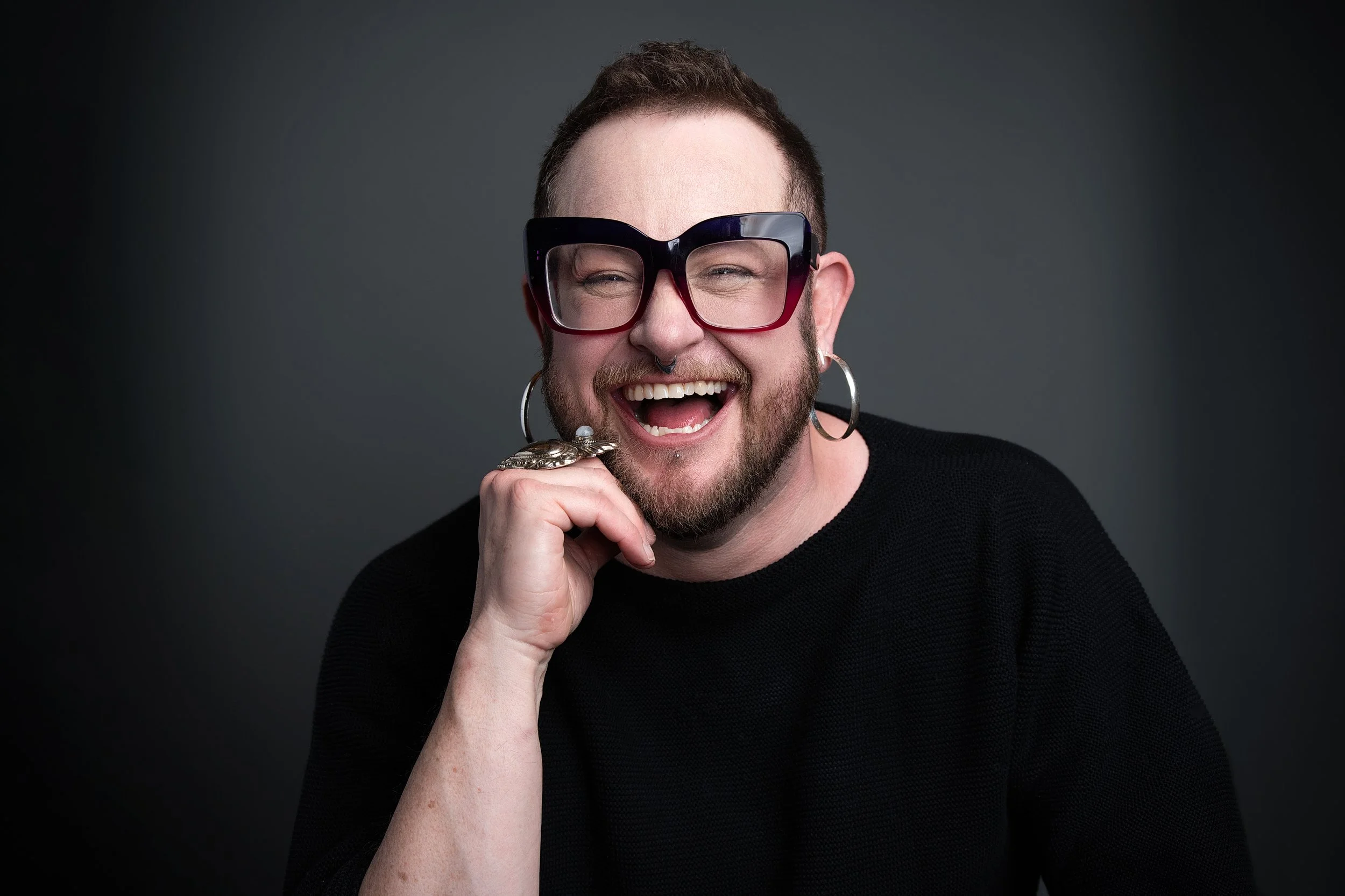 A headshot of Leigh, a light-skinned person with short hair, a beard and facial piercings,in big magenta and black glasses and silver hoop earrings, laughing
