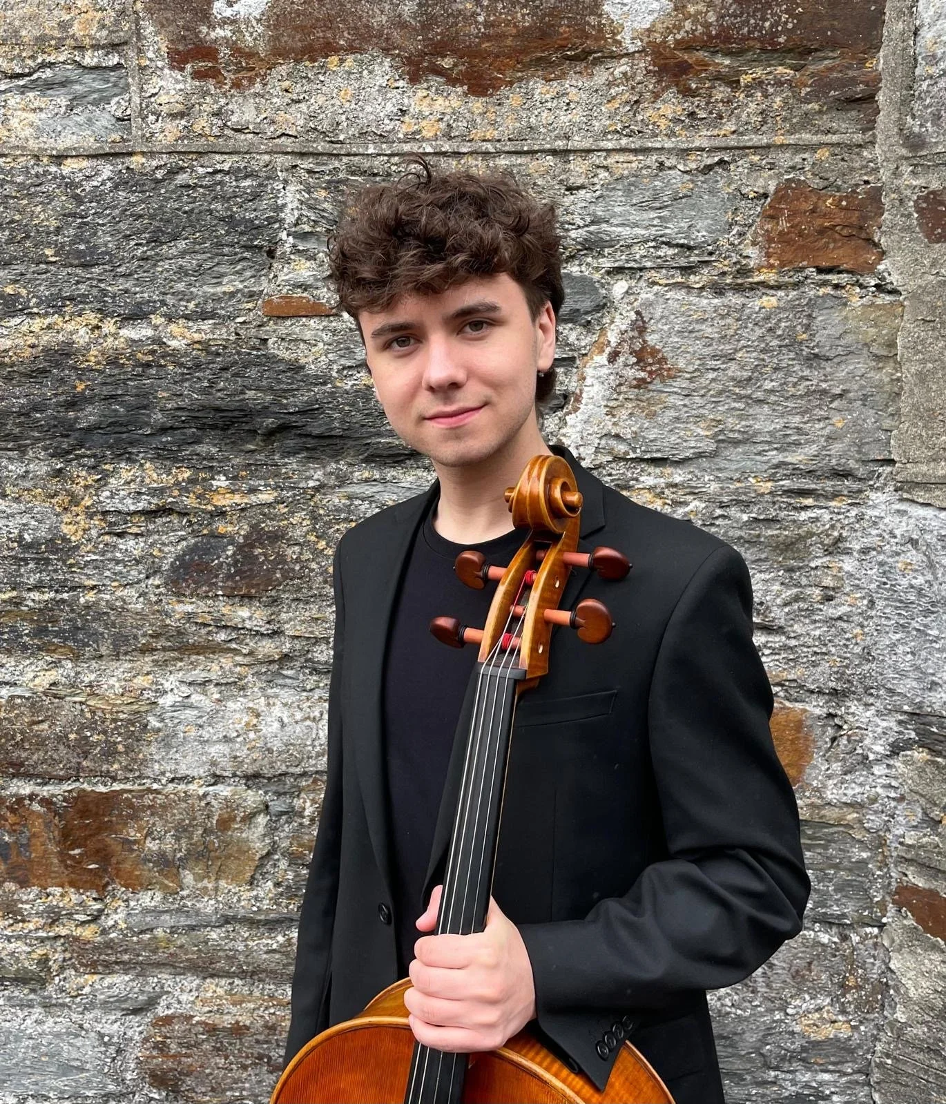 A young man with curly brown hair and fair skin, wearing a black blazer and t-shirt, holding a cello, standing in front of a textured stone wall.