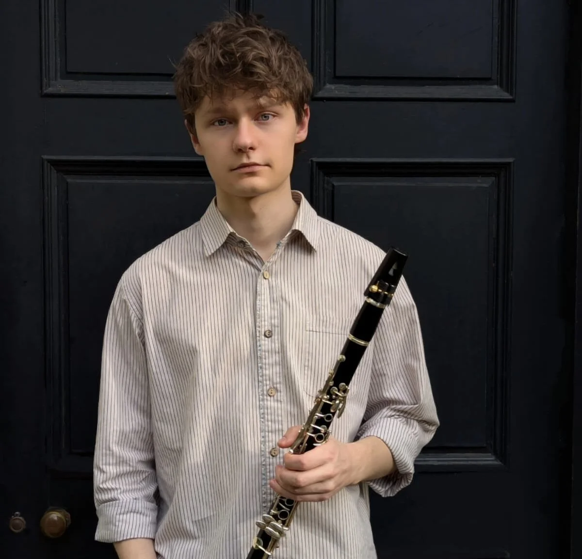 Young man with curly brown hair holding a clarinet, standing in front of a black door.