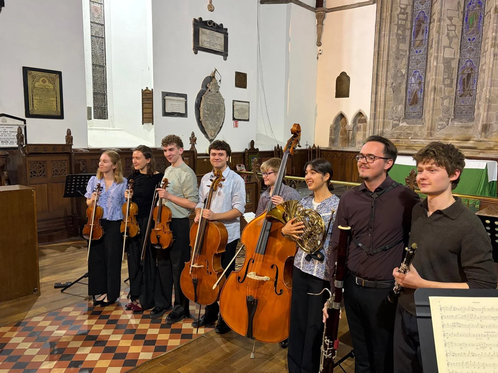 Group of eight musicians with string and wind instruments in St. Flannan's Cathedral, standing on a patterned tile floor, smiling after a performance.