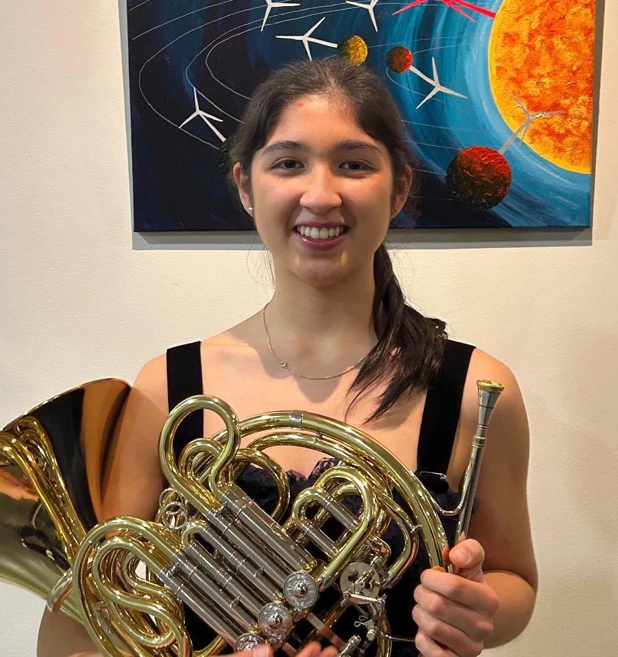 A young woman smiling and holding a French horn, standing in front of a colorful abstract painting of planets and space.